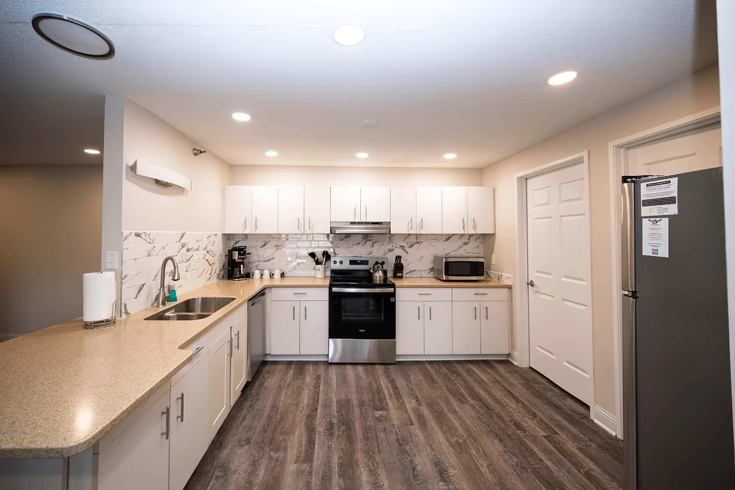 Modern kitchen featuring white cabinets, a marble backsplash, stainless steel appliances, and a center island with a sink. Brightly lit with recessed ceiling lights, the space is designed with a neutral color palette and wood-like flooring. Includes a refrigerator, microwave, and coffee maker.