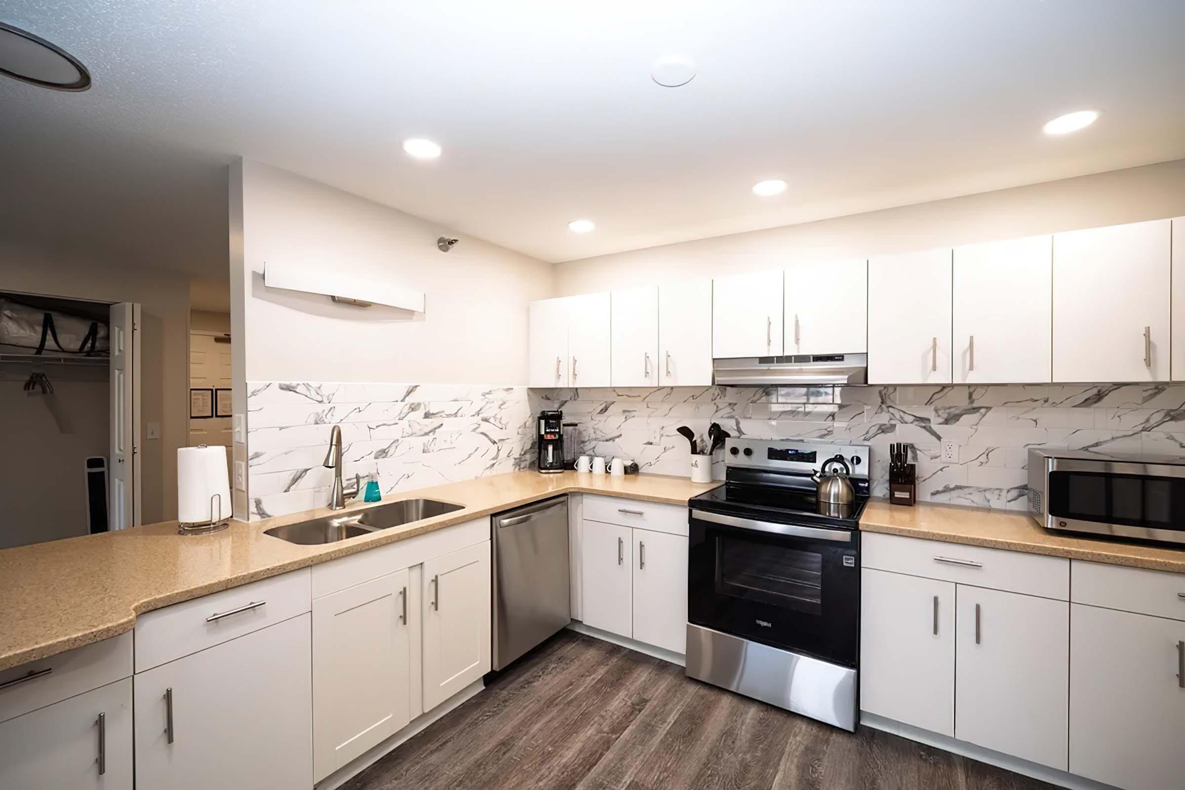Modern kitchen featuring sleek white cabinetry, a marble backsplash, stainless steel appliances including an oven and dishwasher, and a countertop with a sink. There is a coffee maker and a microwave on the countertop, along with a couple of white mugs. The space is well-lit with recessed lighting.