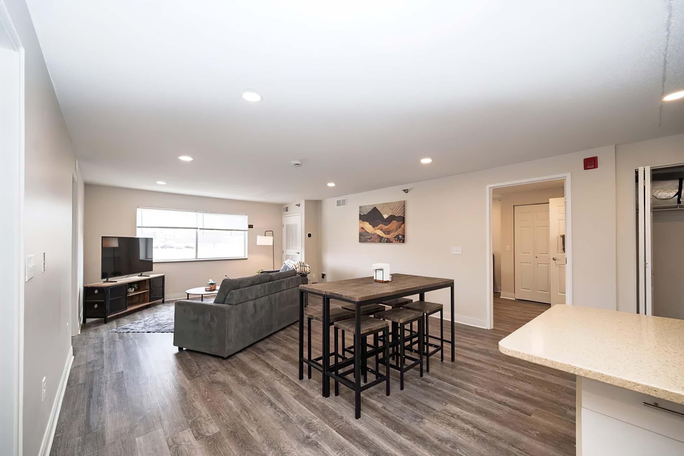 A modern living room and dining area with a gray sofa, wooden dining table with black chairs, and a television. Large windows provide natural light. The walls are painted in neutral tones, and there's a piece of wall art depicting a landscape. A doorway leads to another room.
