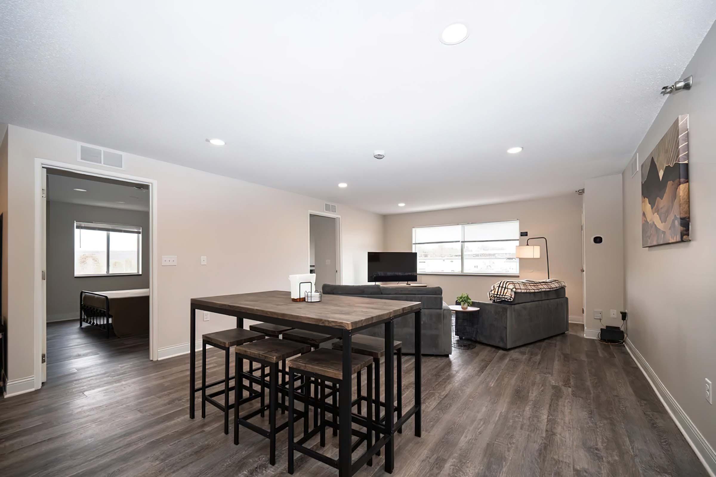 A spacious, modern living area featuring a dining table with stools, a gray sofa, and a television. The room has neutral-colored walls and hardwood flooring, with natural light streaming in through large windows. There is a separate room visible in the background.