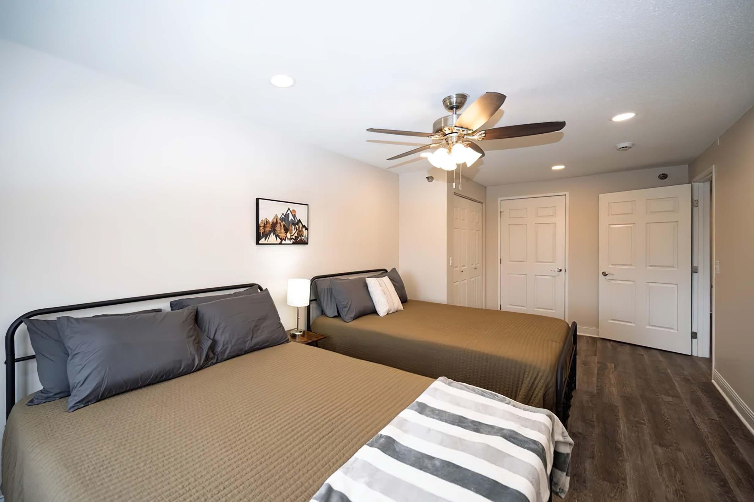 A spacious and modern bedroom featuring two beds with dark gray bedding, a ceiling fan, and a light bedside lamp. The walls are painted in a light color, and a framed picture decorates one wall. Two doors lead to closets or bathrooms, and the flooring is a stylish dark wood.