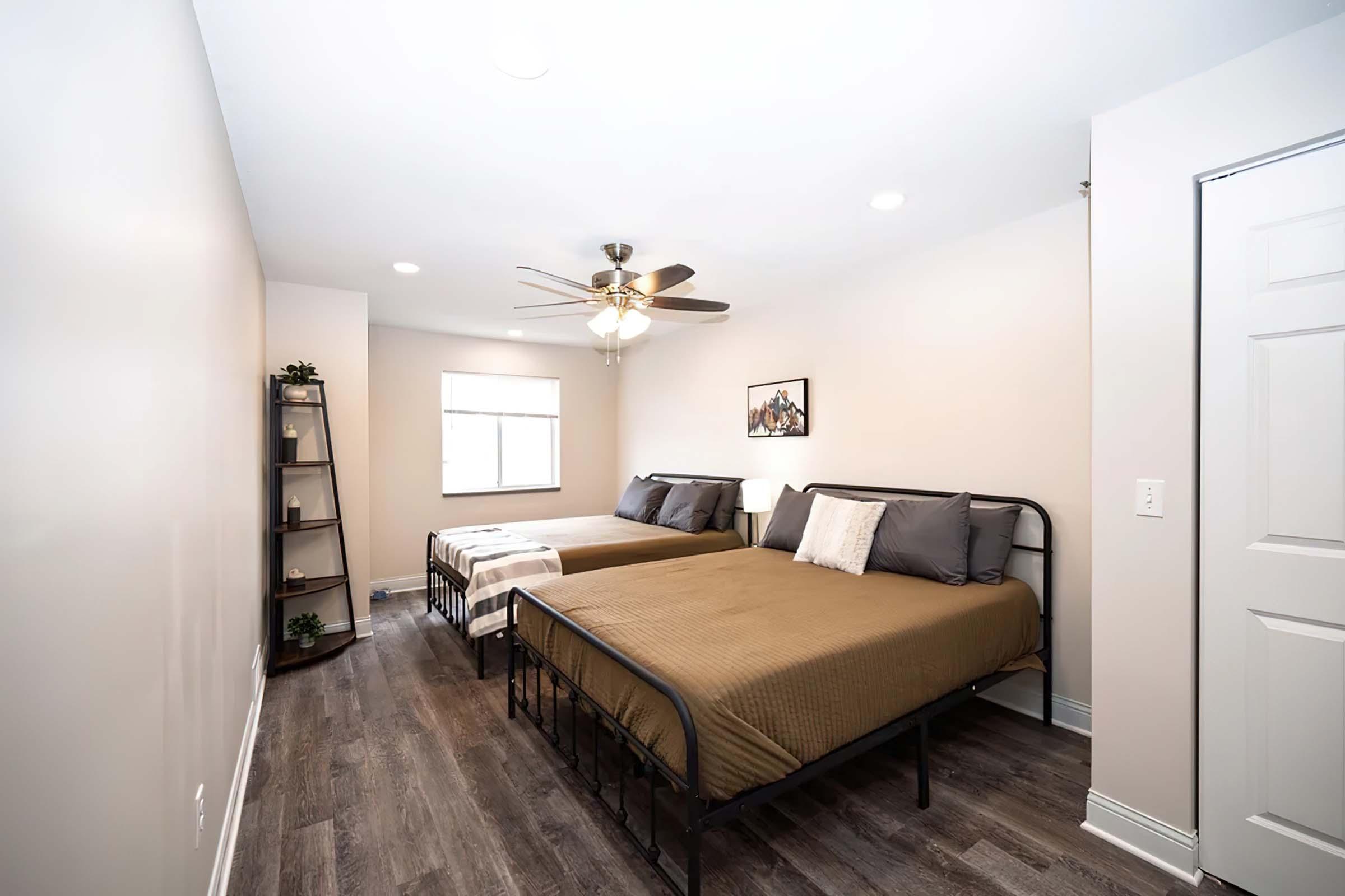 A well-lit bedroom featuring two beds with brown bedspreads. There's a ceiling fan above and a window letting in natural light. A small bookshelf is in the corner, and a door is visible on the right side. The walls are painted a light color, creating a calm atmosphere.