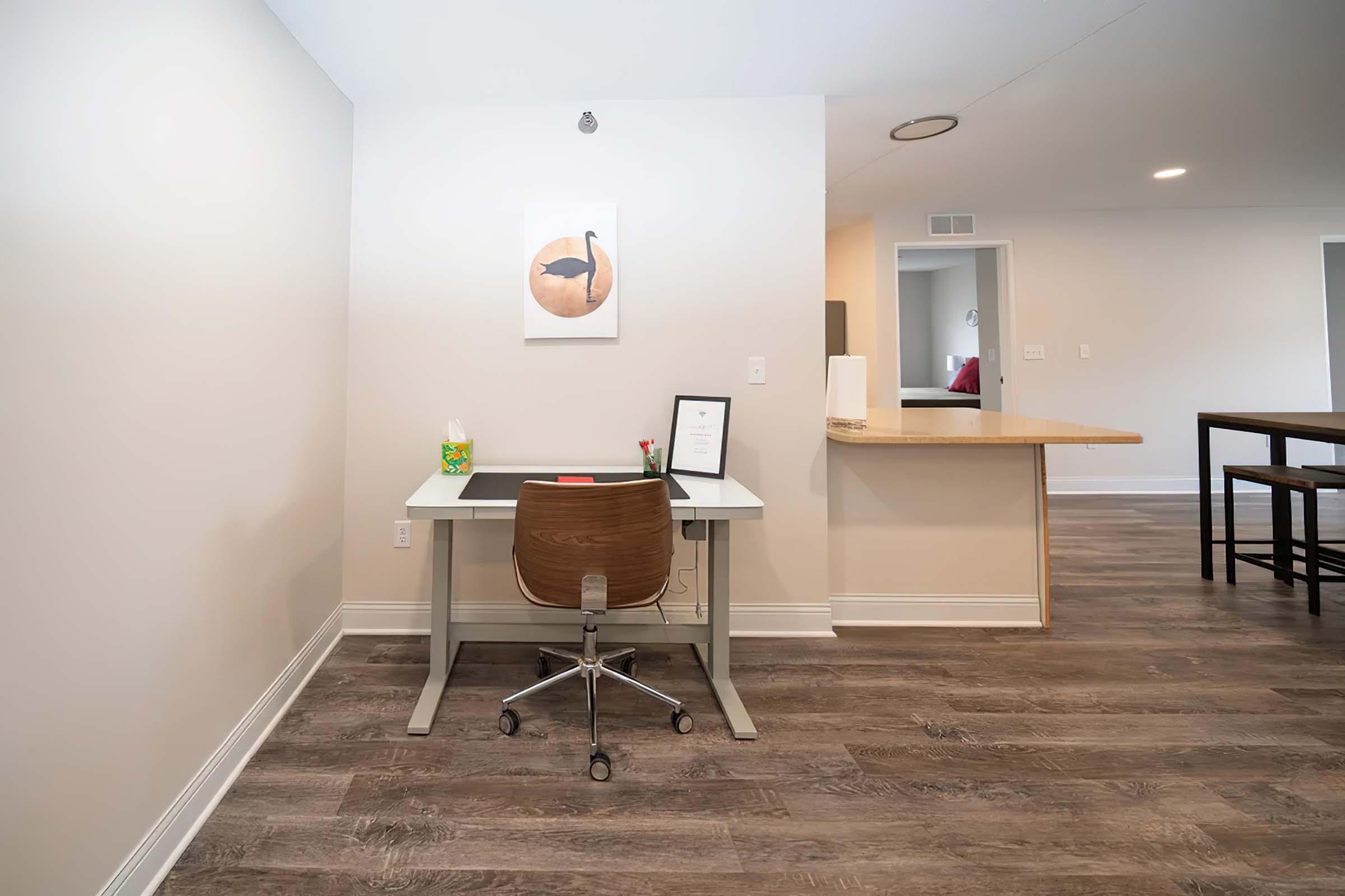 A modern home office setup featuring a small desk with a chair, an award or certificate framed on the desk, and a flower vase. The room has light-colored walls and hardwood flooring, with an open kitchen area visible in the background. Minimalist decor creates a clean and inviting atmosphere.