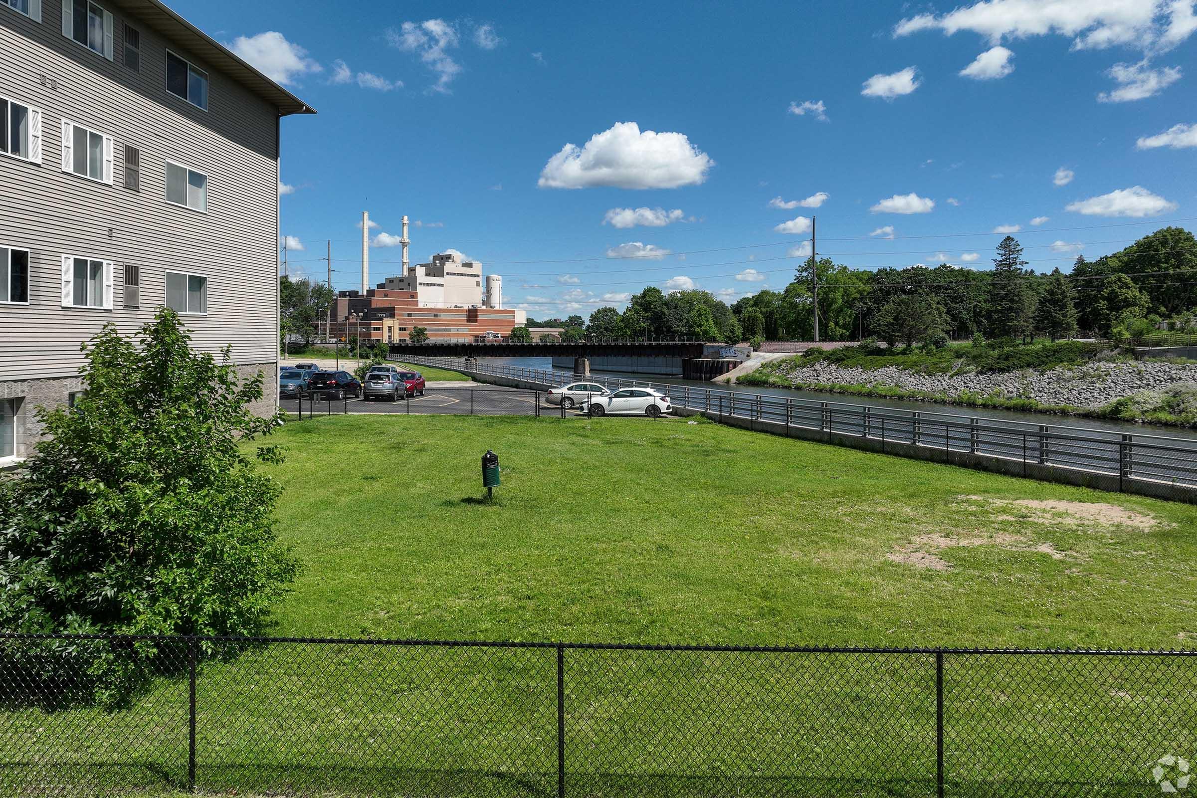 A green grassy area surrounded by a black fence, with a single person standing in the middle. In the background, there are several parked cars and a building near a river. The sky is clear with fluffy white clouds.