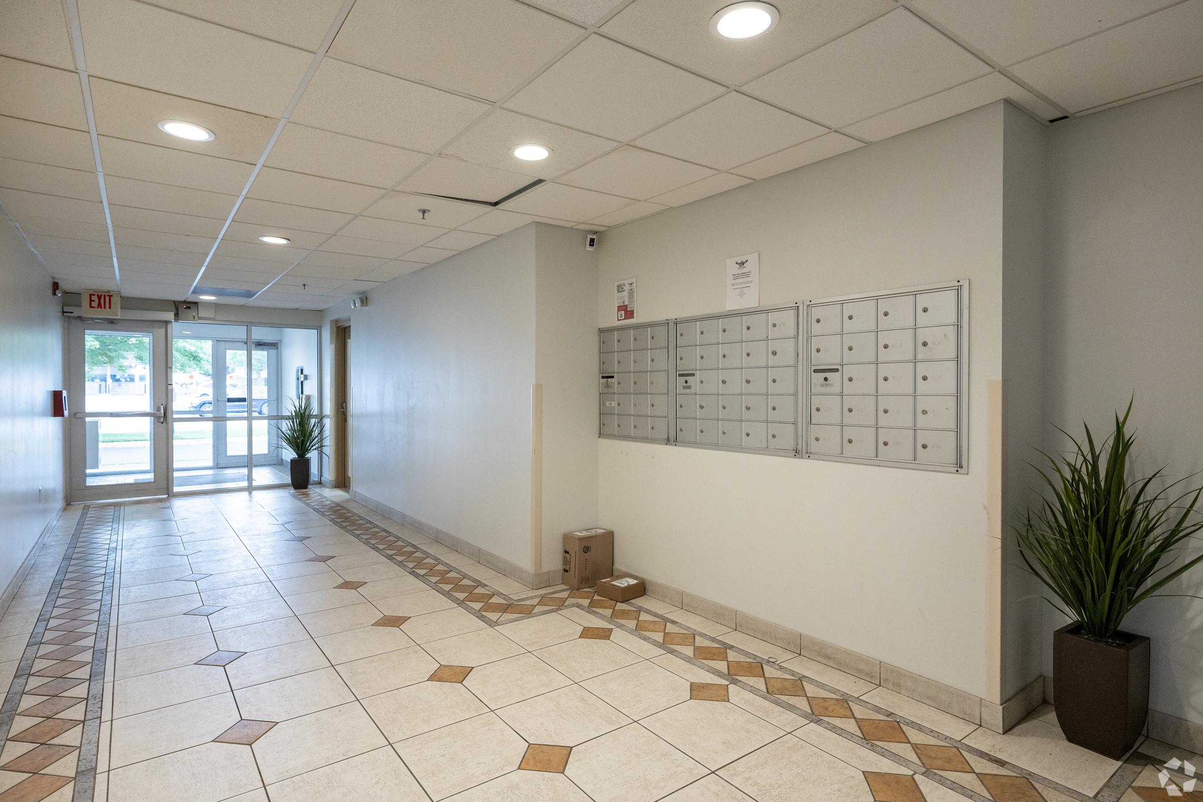 A well-lit corridor featuring mailboxes mounted on a wall. The floor is tiled with decorative patterns, and there are potted plants on either side. At the end of the hallway, glass doors lead outside, creating a welcoming entrance.