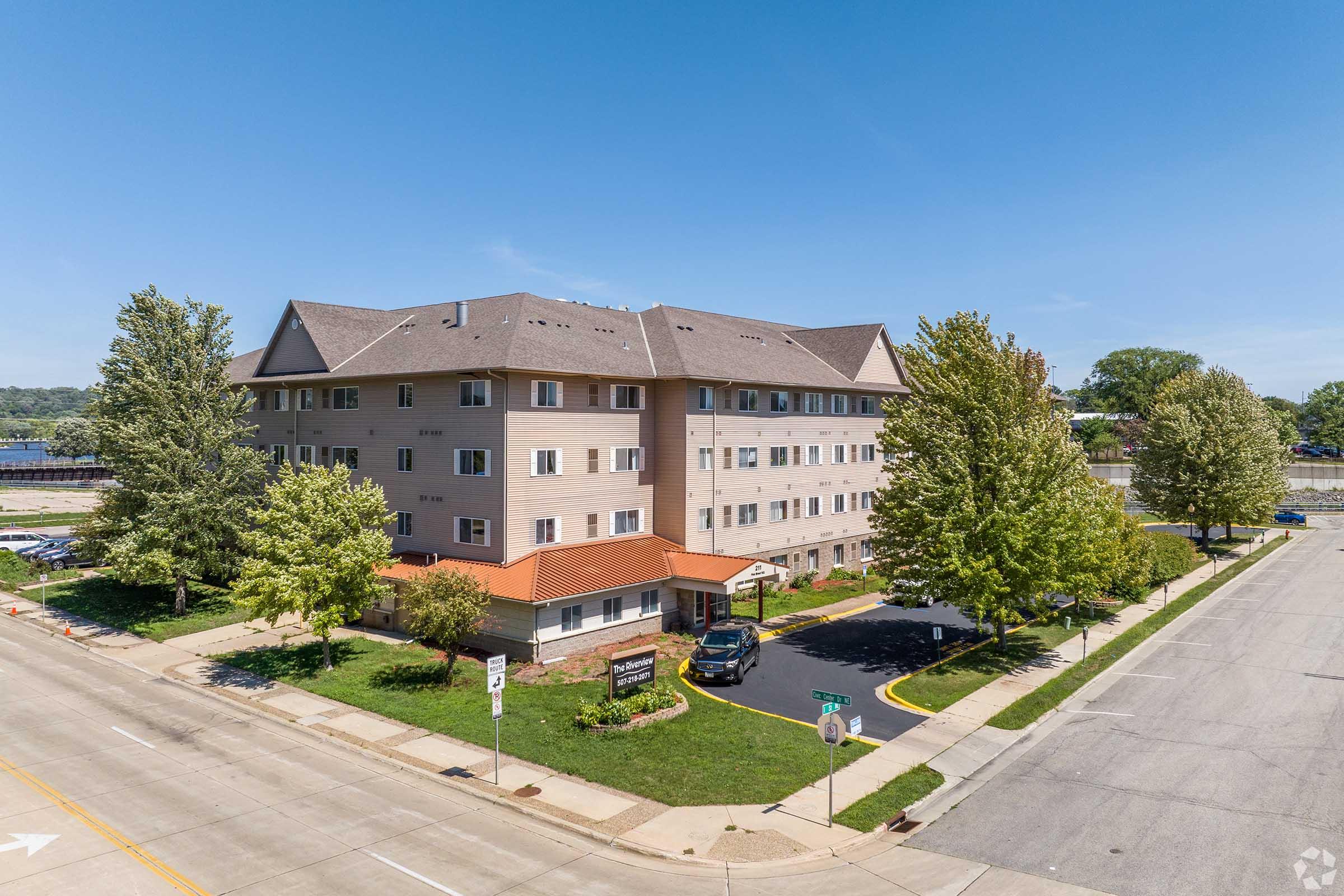 A multi-story residential building with a light-colored exterior and brown roof, surrounded by trees and a well-maintained lawn. Parking spaces are visible in front of the building, and the area features sidewalks along a street on the left side. The sky is clear and blue.