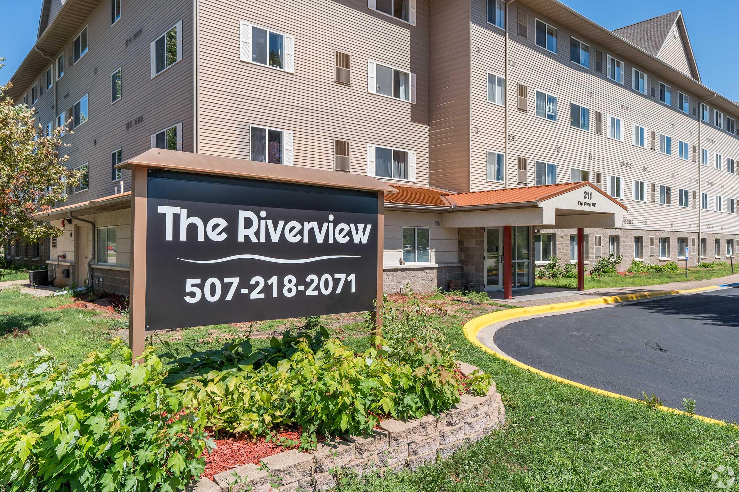 A building exterior featuring a sign that reads "The Riverview" with a contact number, surrounded by greenery and a landscaped entrance. The building has multiple stories with a light-colored facade.