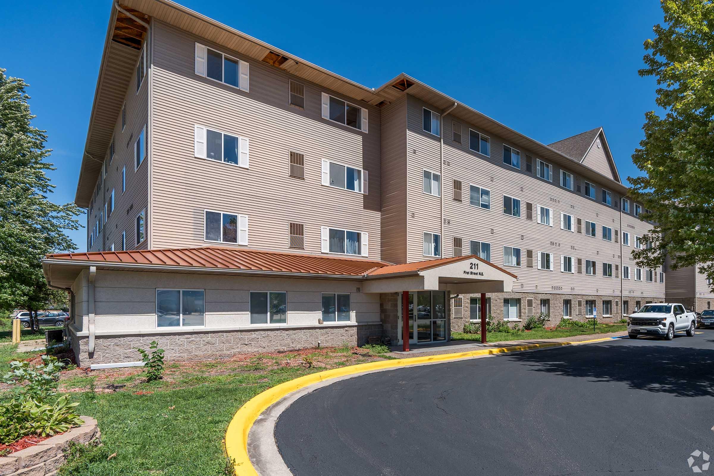 A three-story residential building with a light brown exterior, featuring multiple windows and a peaked roof. The entrance is framed by a covered porch, with landscaped greenery surrounding a curved driveway. Clear blue sky provides a bright backdrop.