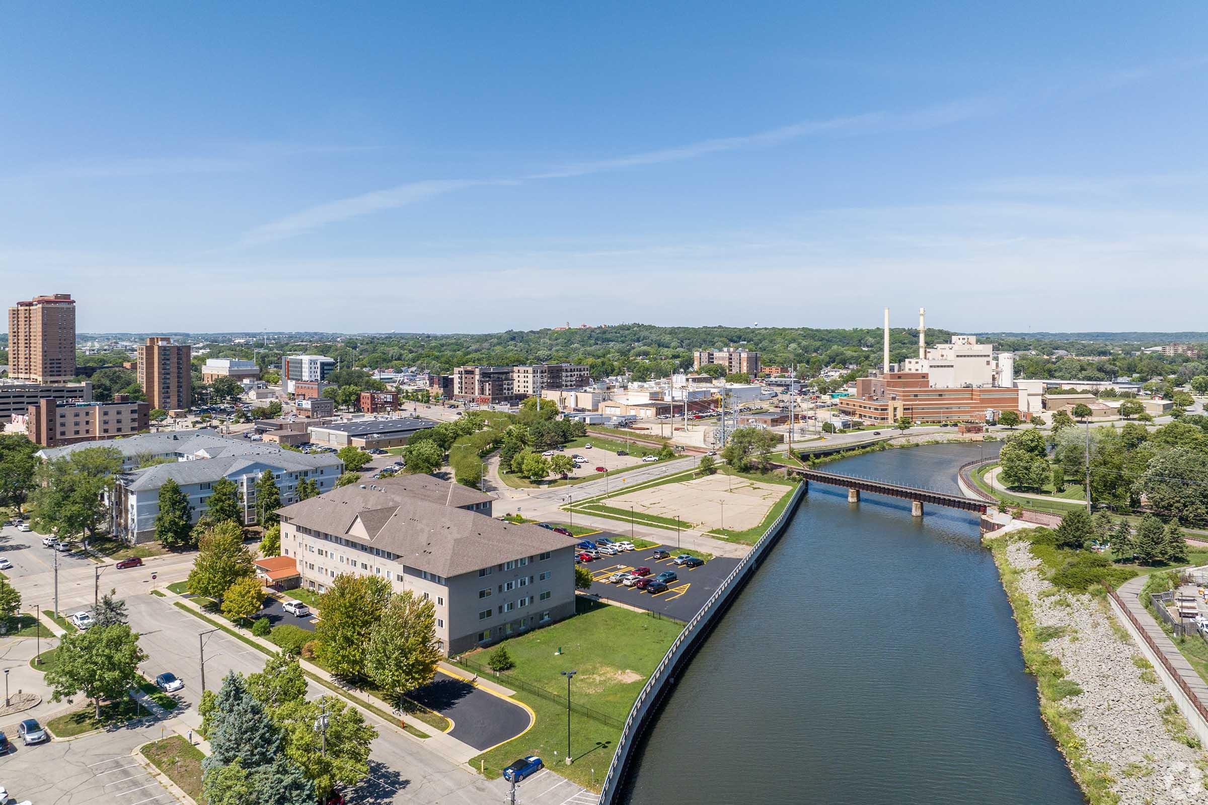 Aerial view of a city featuring modern buildings, a flowing river, and green parks. The skyline showcases a mix of residential and commercial structures, with parking lots and trees lining the streets, set against a clear blue sky.