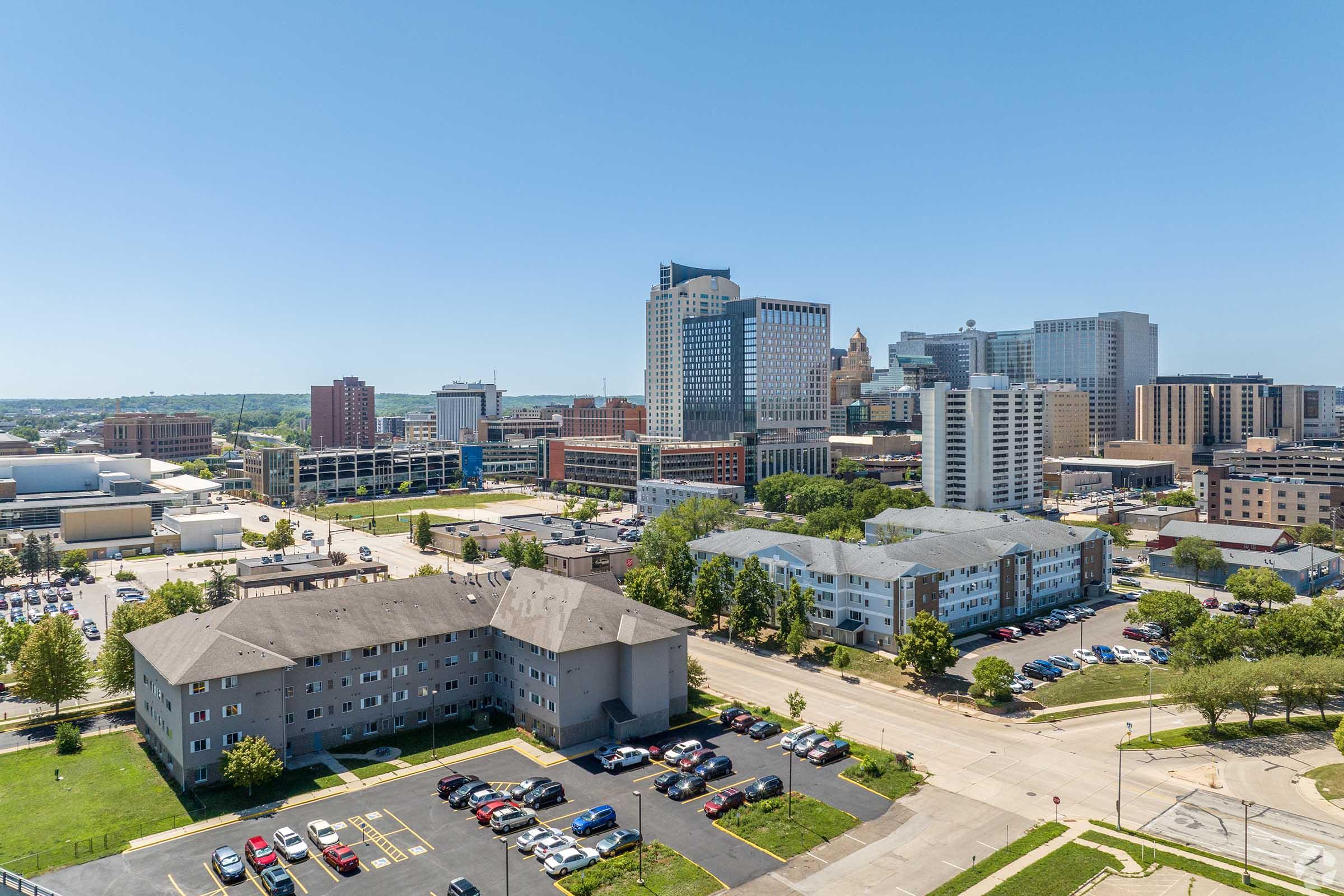 Aerial view of a cityscape featuring modern skyscrapers and commercial buildings, with residential areas and parked cars visible in the foreground. The scene is set under a clear blue sky, showcasing a vibrant urban environment.