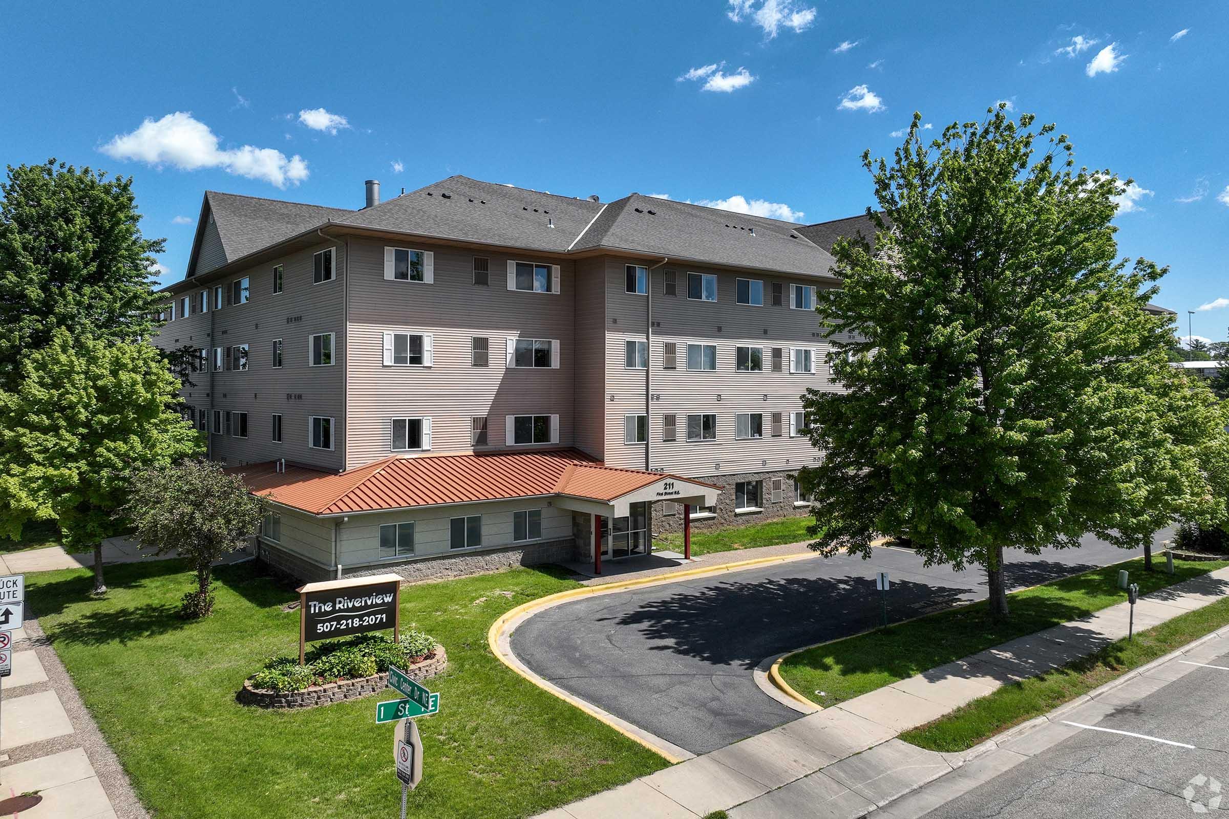 A multi-story building featuring a flat roof and a covered entrance. The exterior is light-colored with a red-tiled roof and surrounded by greenery. The building is situated on a tree-lined street under a clear blue sky. A sign in front indicates the name and contact number of the establishment.