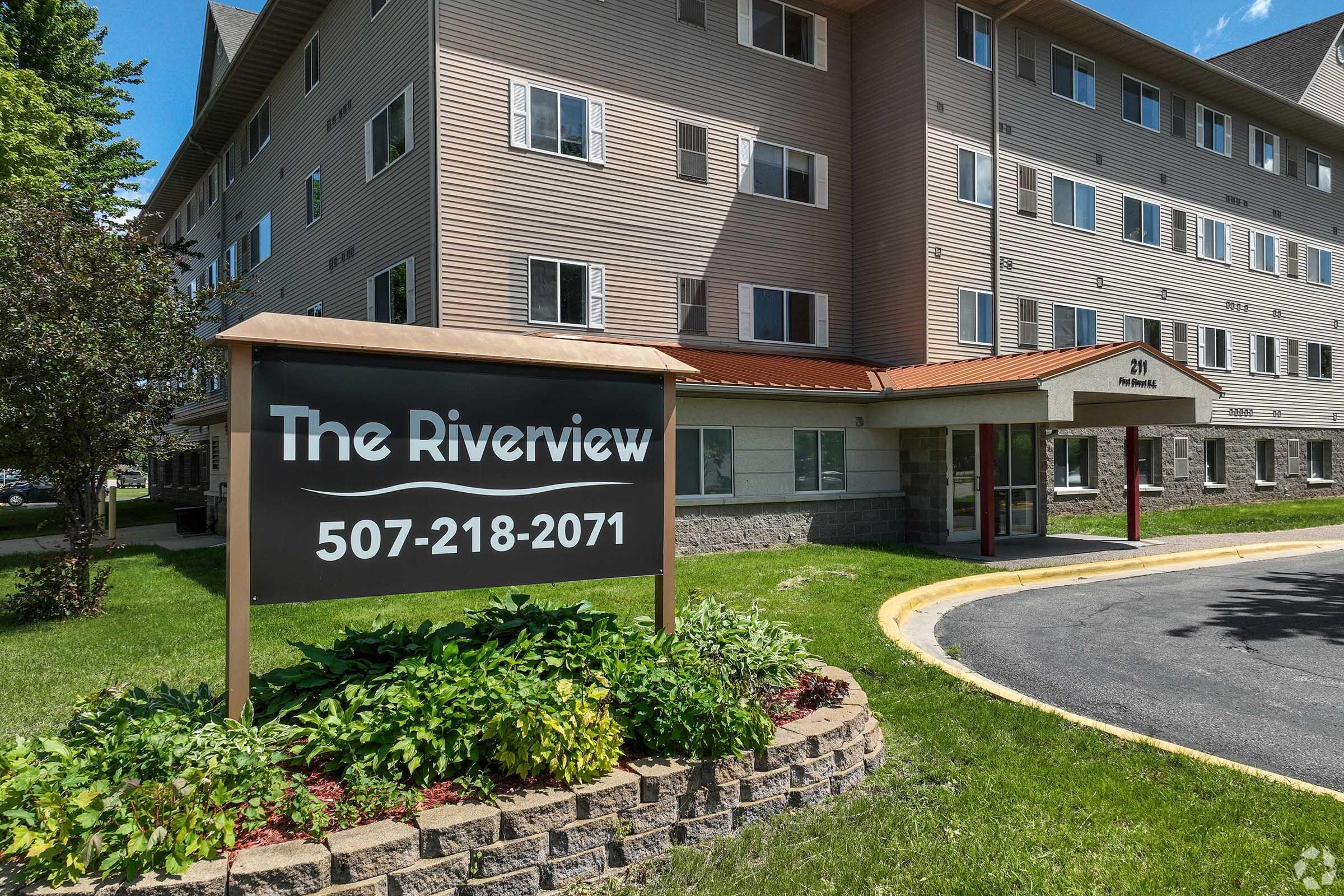 A exterior view of "The Riverview" building, showcasing a multi-story structure with a modern design. A sign at the entrance displays the name and phone number, surrounded by neatly maintained landscaping and grass. The scene is bright and inviting, with blue skies in the background.