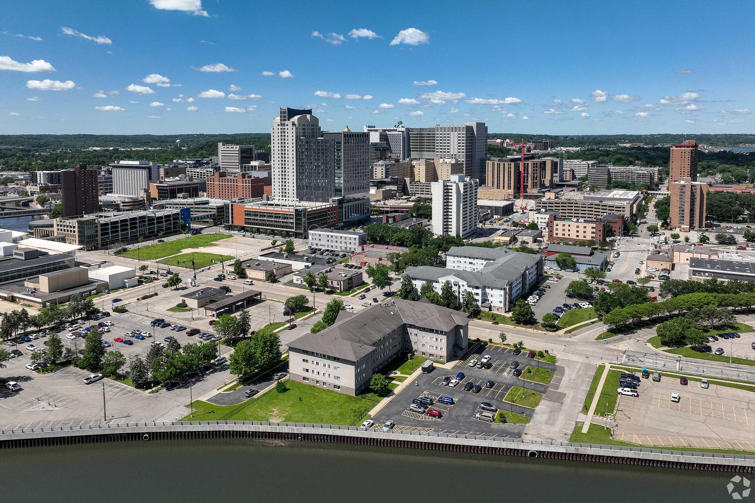 Aerial view of a city skyline featuring various tall buildings, including commercial and residential structures. In the foreground, there are parking lots and green spaces along a waterfront. The sky is partly cloudy, and the landscape extends into the distance with more buildings and greenery visible.