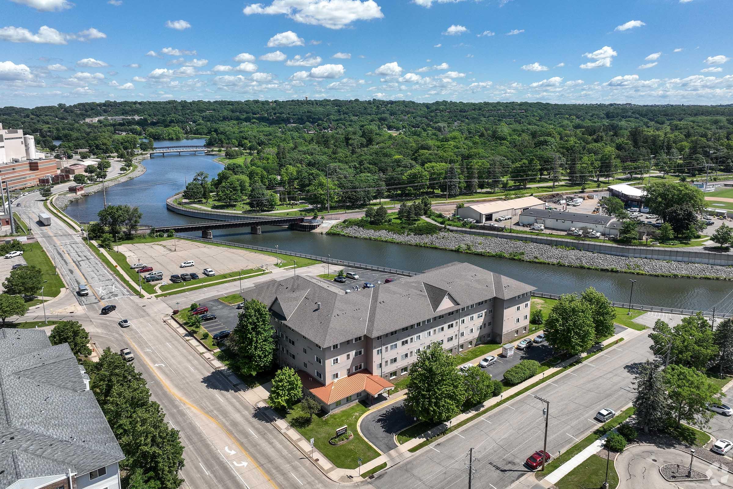 Aerial view of a river running through a green landscape, bordered by trees and buildings. In the foreground, a multi-story building is visible alongside a parking lot. The scene includes a road stretching alongside the river, with scattered vehicles and blue sky dotted with fluffy clouds.