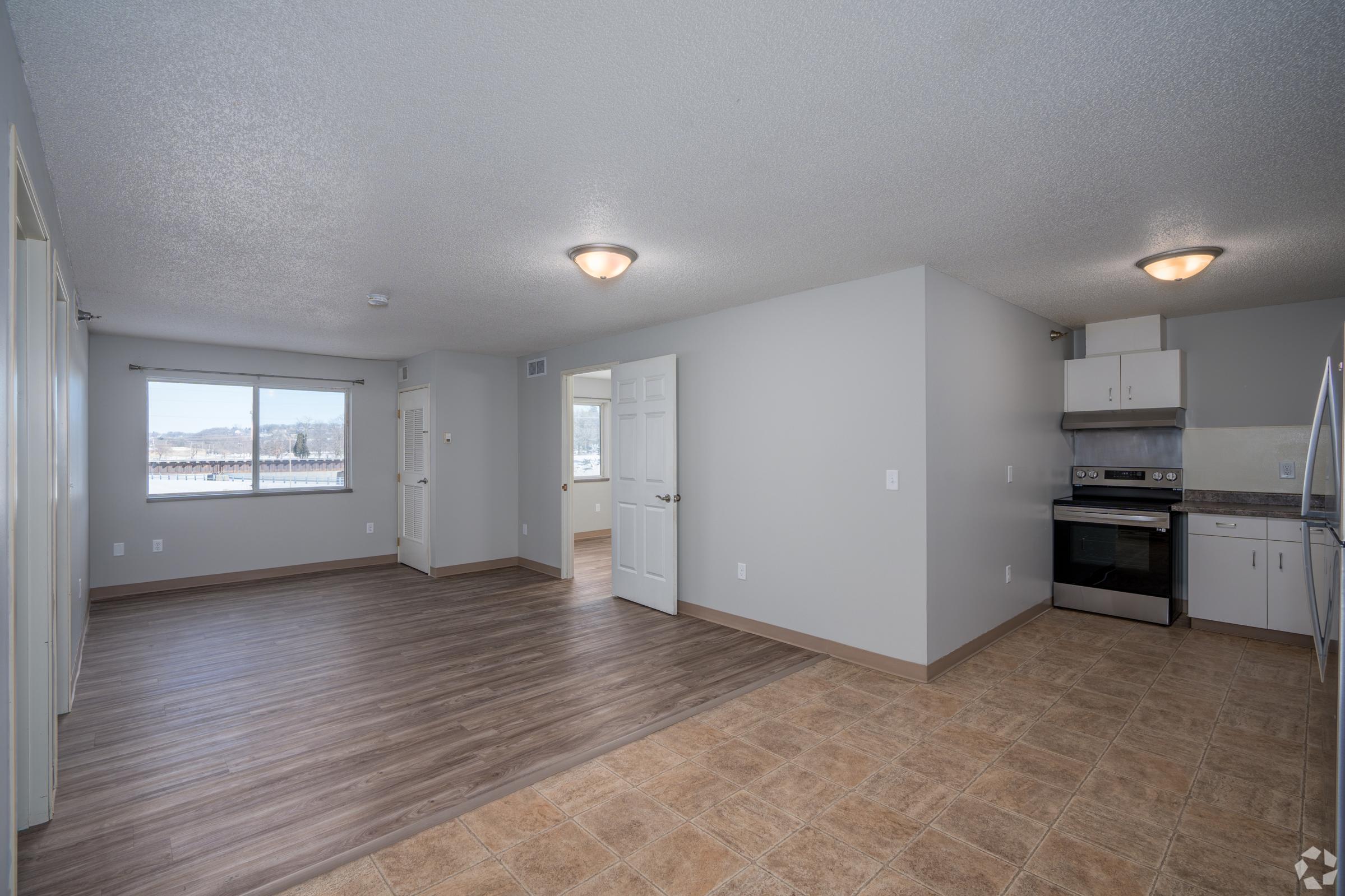 A spacious, newly renovated apartment interior featuring light-colored walls and a combination of laminate and tile flooring. The layout includes a kitchen area with stainless steel appliances, a window providing natural light, and doorways leading to other rooms.