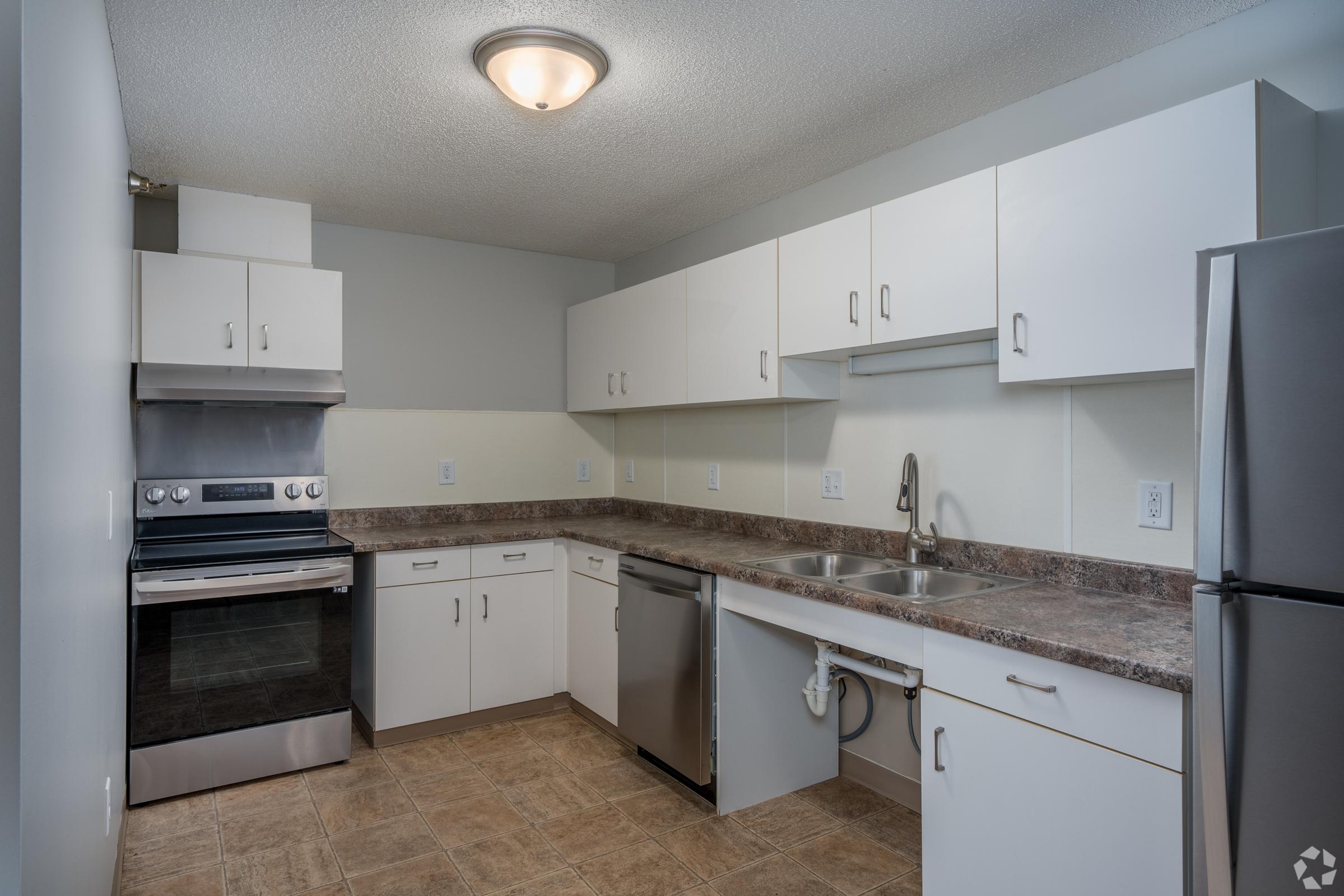 Modern kitchen featuring white cabinetry, a stainless steel refrigerator, an oven with a cooktop, and a double sink. The countertops are a neutral stone color, and the floor consists of tiles. Bright overhead lighting enhances the clean and spacious appearance of the room.