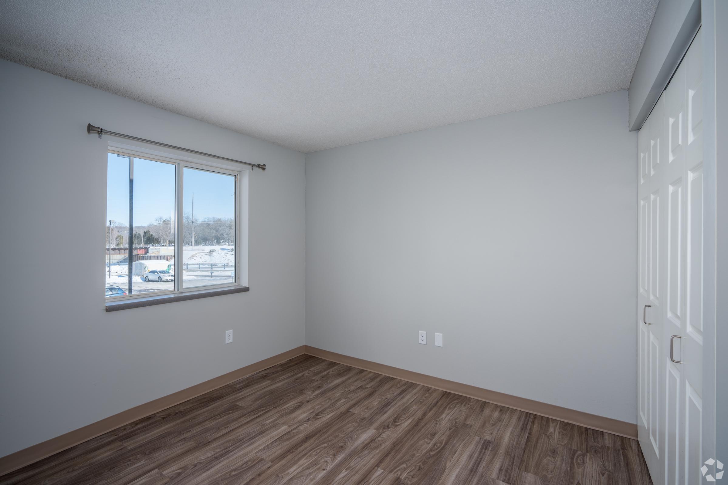 A well-lit, empty bedroom featuring light gray walls and a large window with a view. The floor is made of brown laminate, and there are two white closet doors on one side. The space appears neat and ready for furnishing.