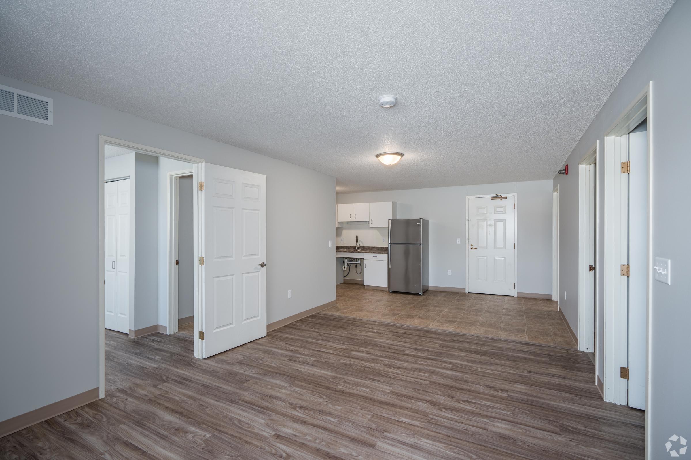 Spacious interior of an apartment with wooden flooring, featuring multiple doors leading to different rooms. A kitchen area is visible in the background, equipped with a stainless steel refrigerator. The walls are painted light gray, and there is a ceiling light fixture providing illumination.