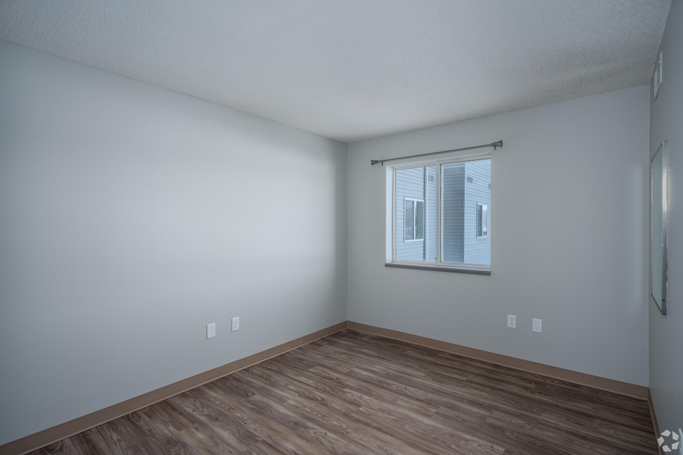 Empty room with light gray walls and wooden flooring. A window with sheer curtains allows natural light to enter, illuminating the space. There are no furnishings or decorations, giving it a clean and minimalist look.