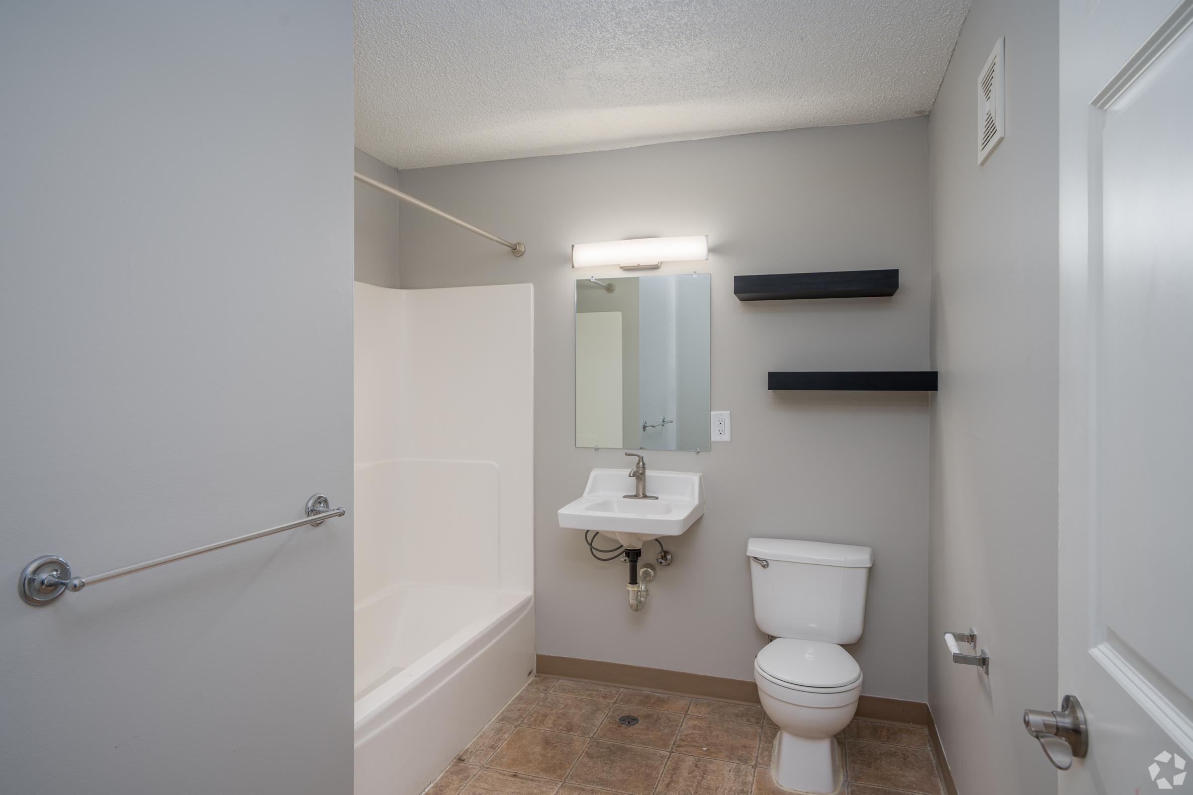 A clean, modern bathroom featuring a white bathtub with a shower, a wall-mounted sink with a mirror above, a toilet, and two wall shelves. The walls are painted light gray, and the floor has tiled squares in a neutral tone.