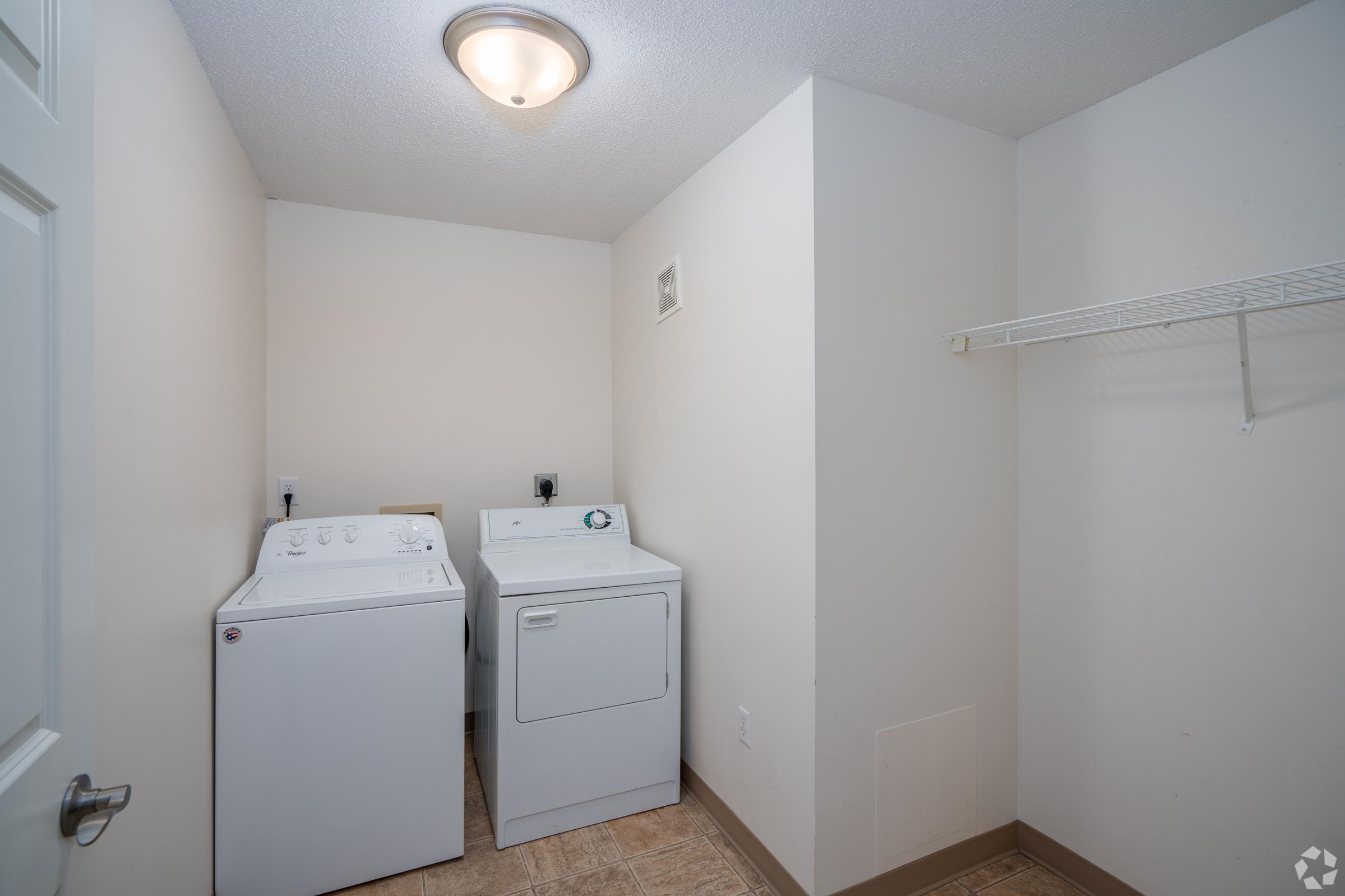 A bright laundry room featuring a white washing machine and dryer side by side. The room has neutral-colored walls and a ceiling light. There is a metal wire shelf mounted on the wall and a small ventilation opening. The floor is tiled, creating a clean and organized space.
