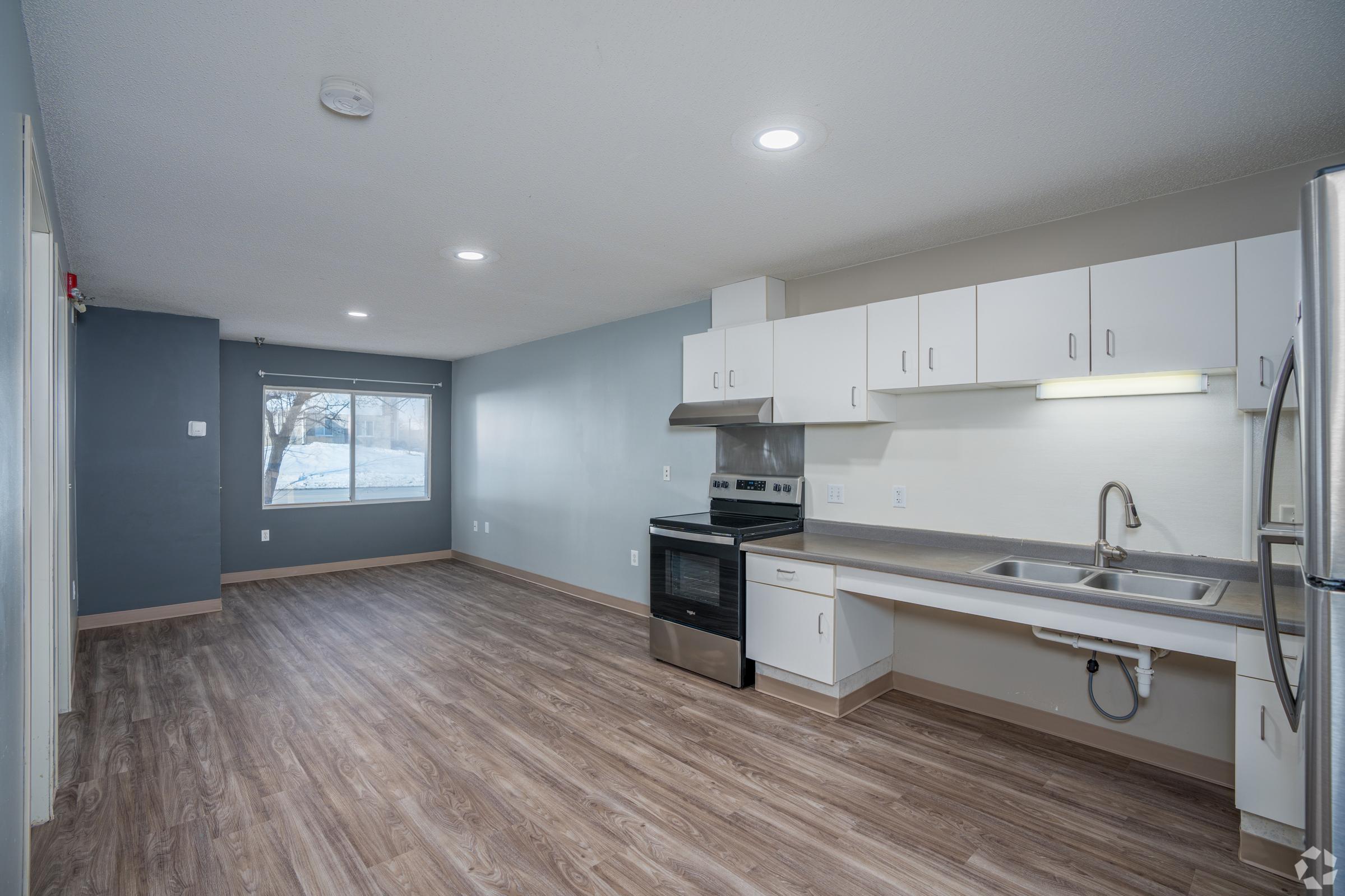 A spacious kitchen and living area with modern fixtures. The kitchen features a stainless steel oven, sink, and white cabinets. Large window allows natural light, while vinyl flooring adds warmth. The walls are painted in soft colors, providing a clean and inviting atmosphere.