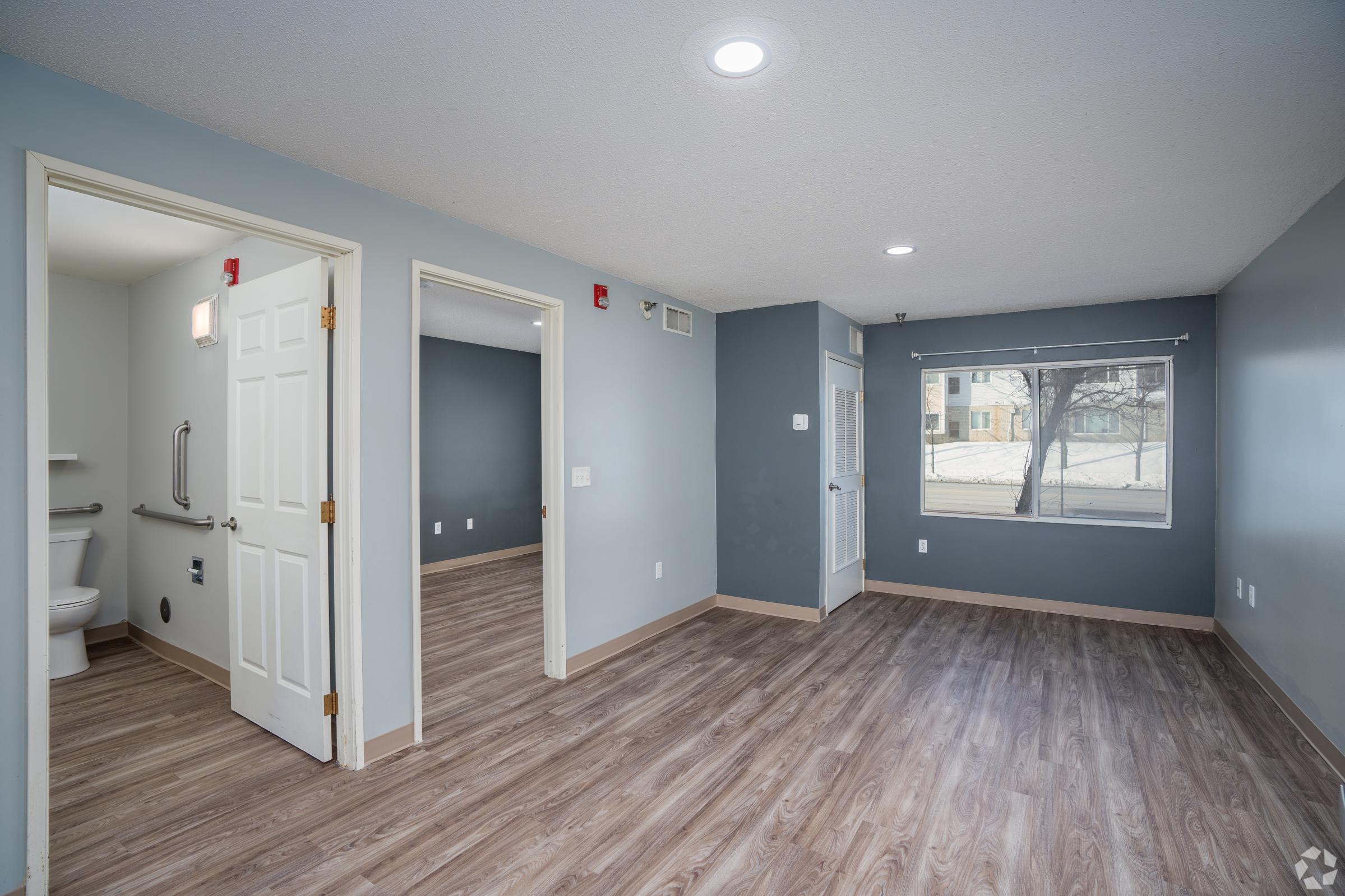 Interior view of a modern room featuring light blue walls, wooden flooring, a window with a view outside, and an adjoining bathroom area with grab bars. Two doors lead to different areas, showcasing a spacious, well-lit layout suitable for living or working.