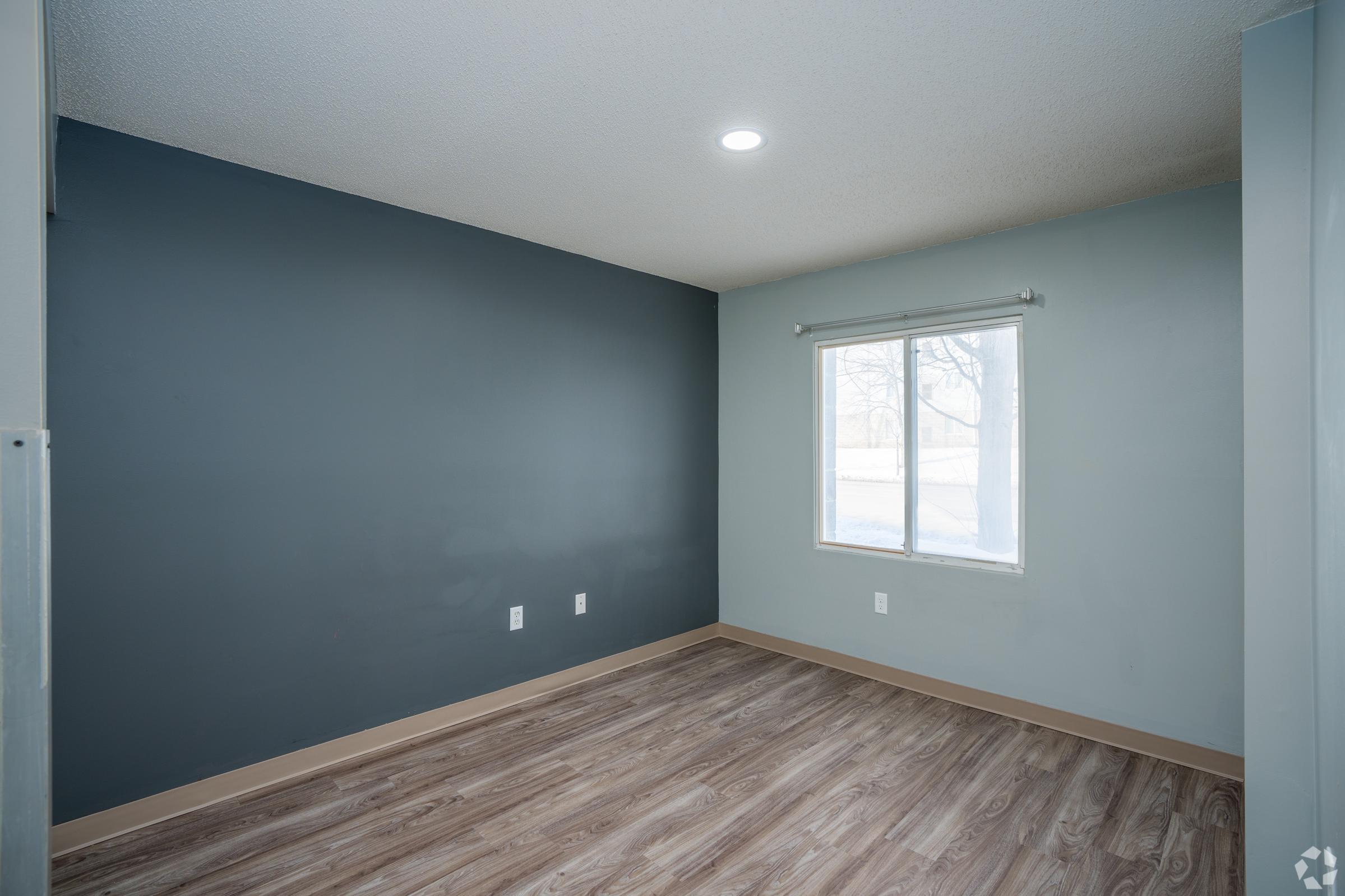 Empty room with gray walls and a large window letting in natural light. The flooring is a light wooden style, and there are two electrical outlets visible on one wall. A bright ceiling light illuminates the space, creating a clean and airy atmosphere.