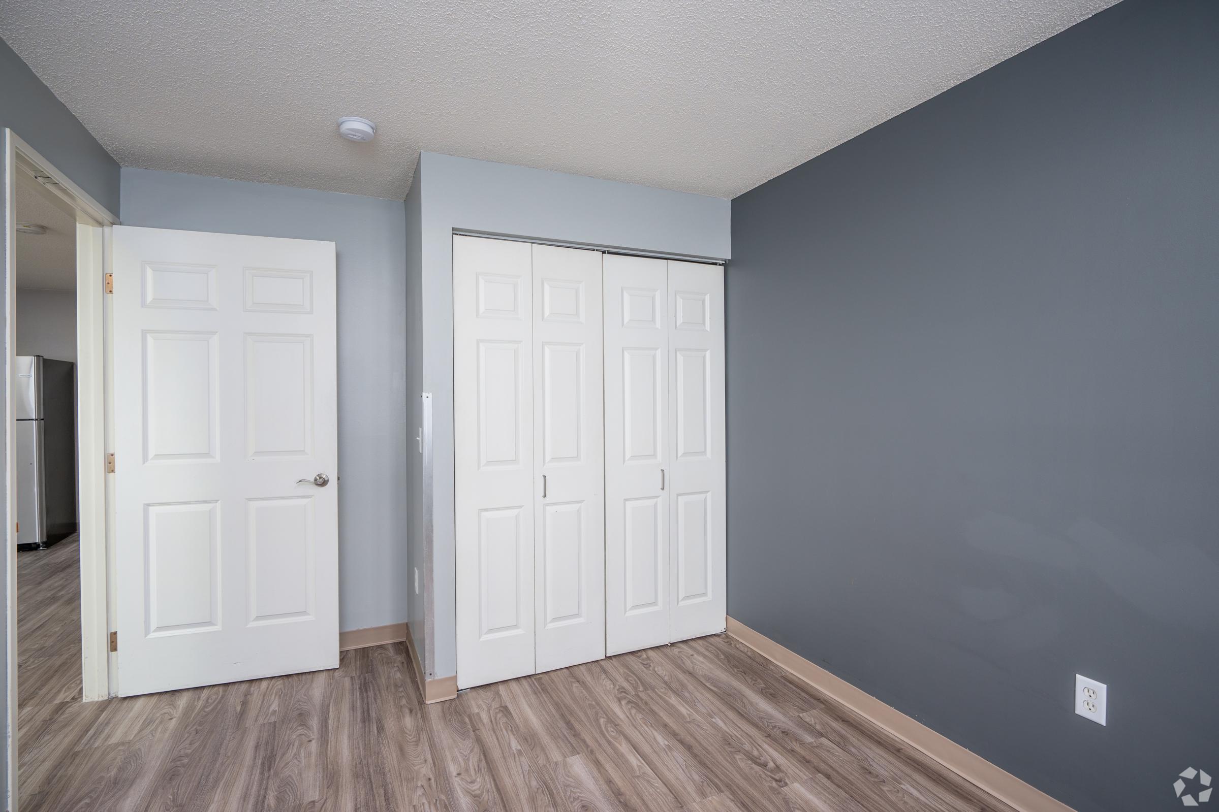 A corner of a room featuring a gray wall, a closet with white bi-fold doors, and light-colored laminate flooring. A door leads to another area, and a glimpse of a kitchen can be seen in the background. The space appears bright and well-maintained, suitable for living or storage.