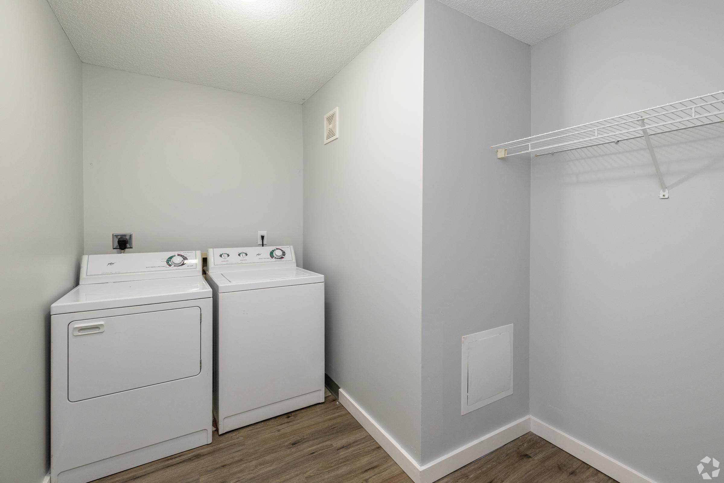 A laundry room featuring two white appliances: a washing machine and a dryer. The walls are painted light gray, and a wire shelf is mounted above the appliances. The floor is wooden, and there’s a small vent or access panel on the wall. The room is well-lit and uncluttered.