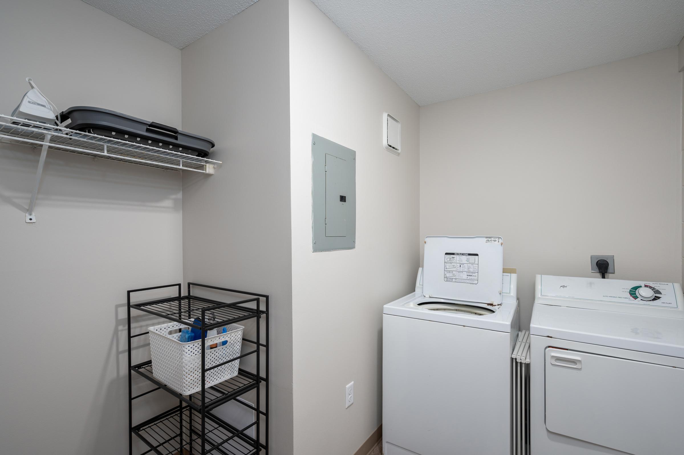 A clean, organized laundry room featuring a stacked washing machine and dryer, a wire shelf above with a few items, and a black metal shelving unit with a storage basket. The walls are painted in a light color, and there is an electrical panel on the wall.