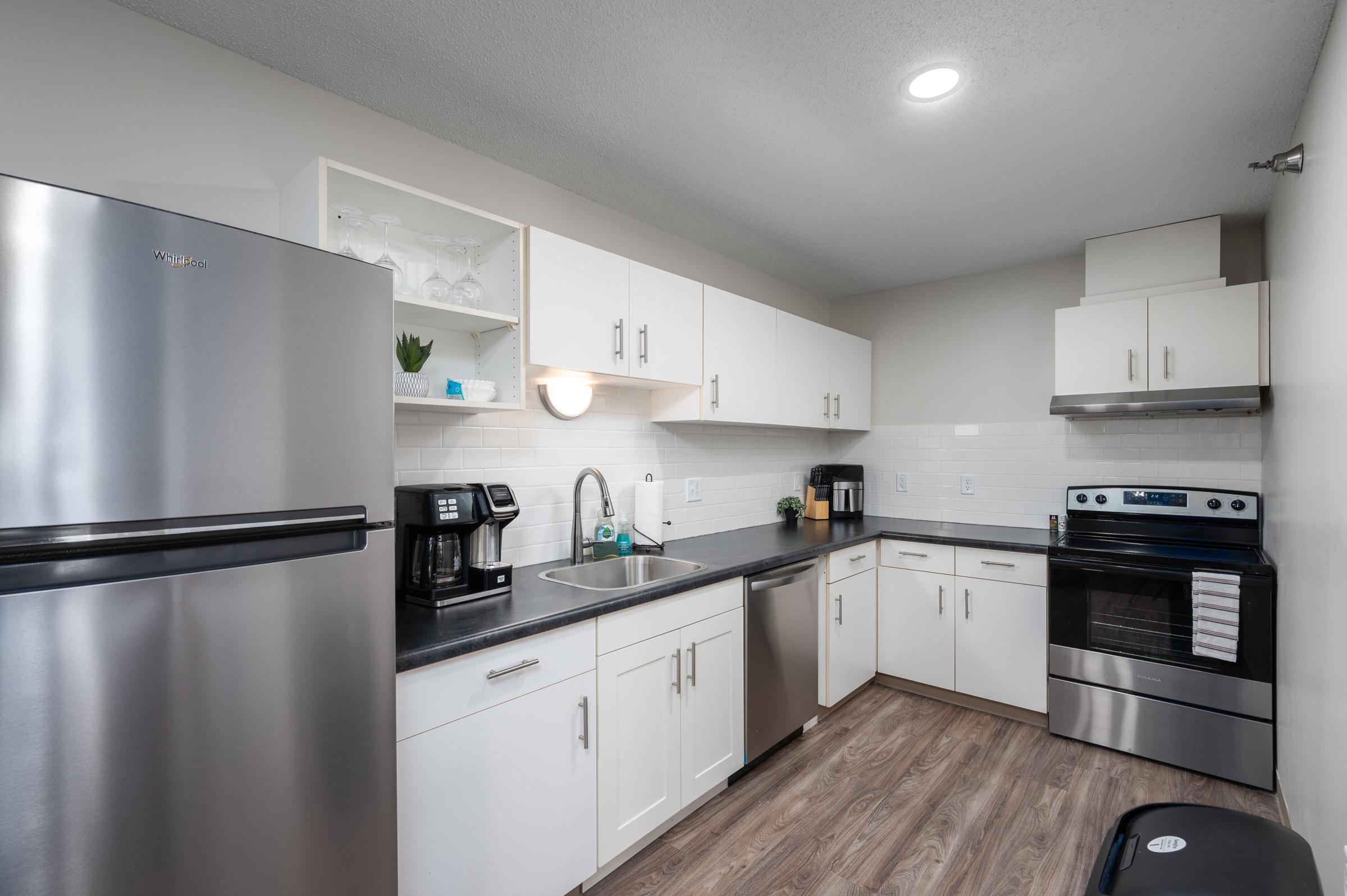 Modern kitchen featuring stainless steel appliances, including a refrigerator and oven. There are white cabinets, a black countertop, and a stainless steel sink. Natural light from overhead lighting illuminates the space, complemented by decorative items on the shelves. The flooring is a light wood finish.