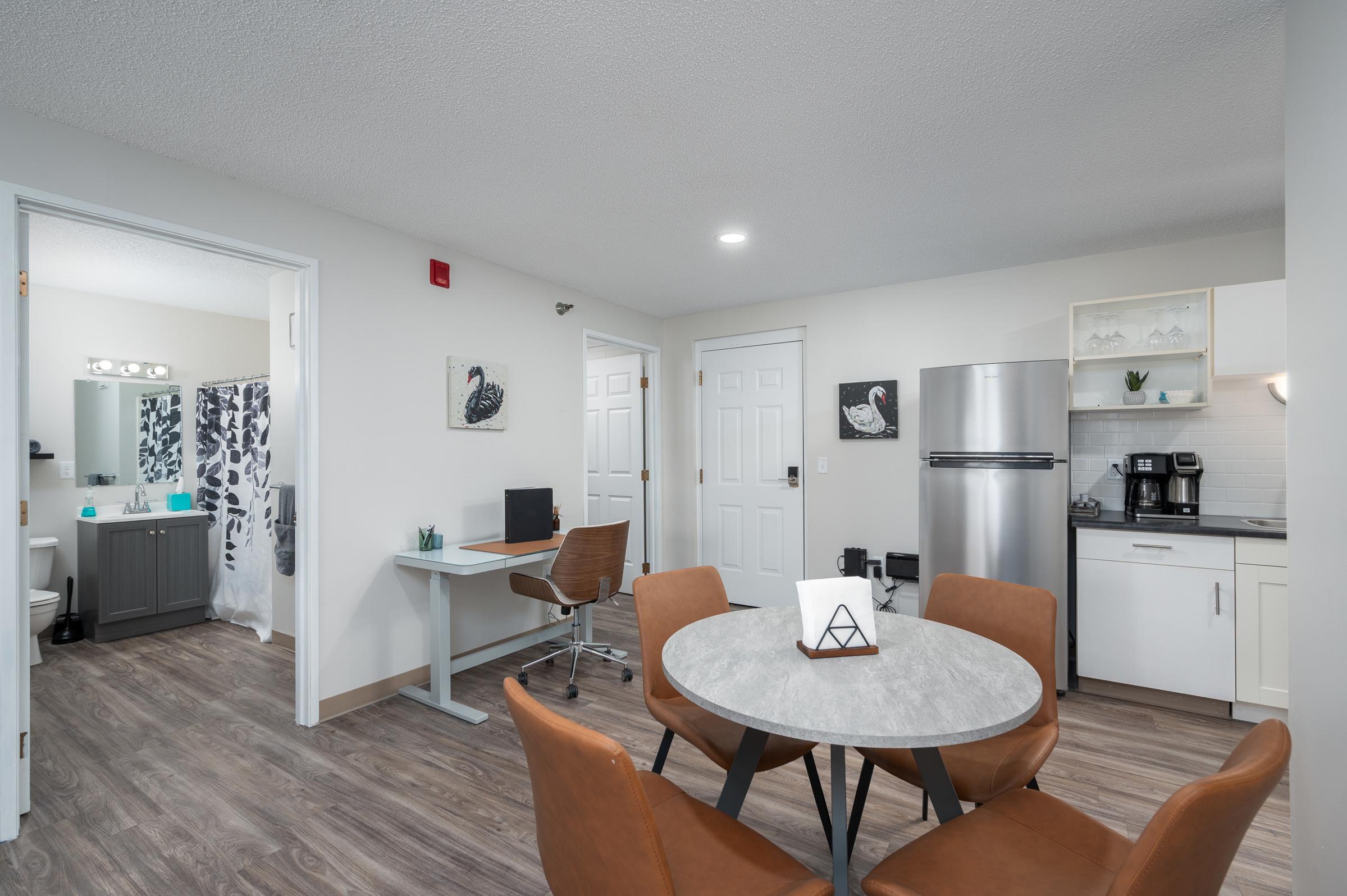 Modern apartment interior featuring a small dining area with a round table and chairs, a kitchen with stainless steel appliances, and a study desk. A bathroom is visible through an open door, and decorative elements like plants and art add to the contemporary decor.