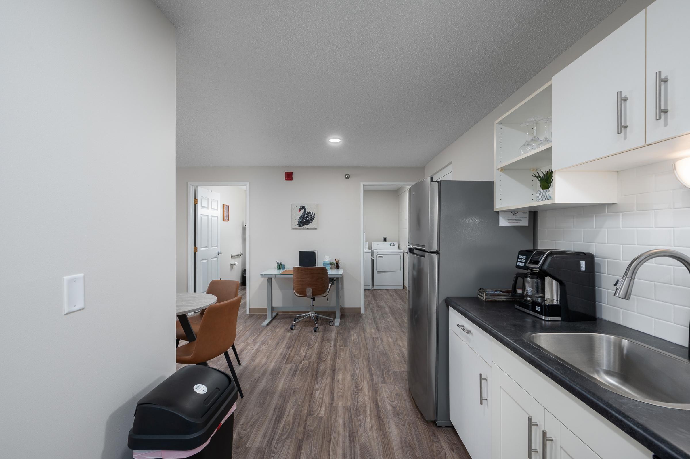 A modern kitchen and dining area featuring a refrigerator, sink, countertop, and coffee maker. There is a small dining table with two chairs, an office desk with a computer in an adjacent room, and a laundry area visible in the background. The flooring is laminate wood, and the walls are light-colored.
