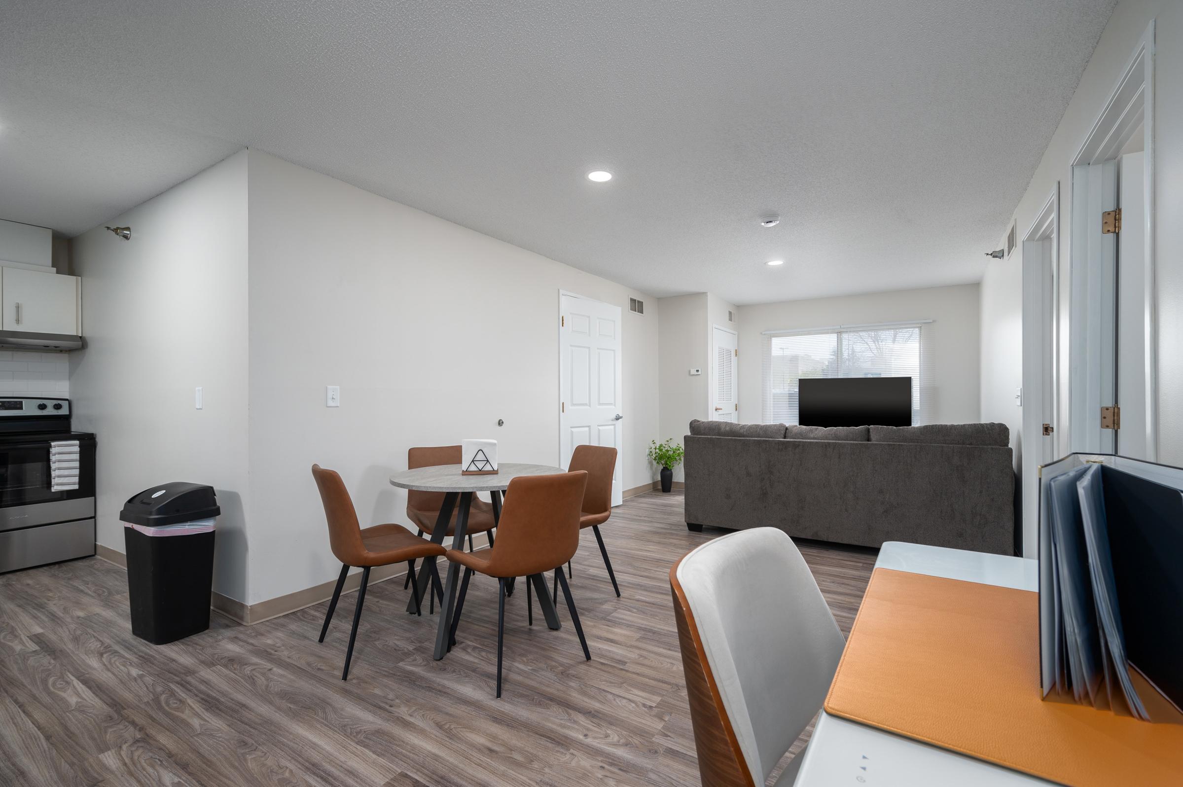 Modern living space featuring an open layout. A dining table with four brown chairs is set beside a gray couch and a wall-mounted television. A small desk with an orange folder is visible, along with a trash can. The walls are light and the flooring is wood-like, creating a bright, inviting atmosphere.