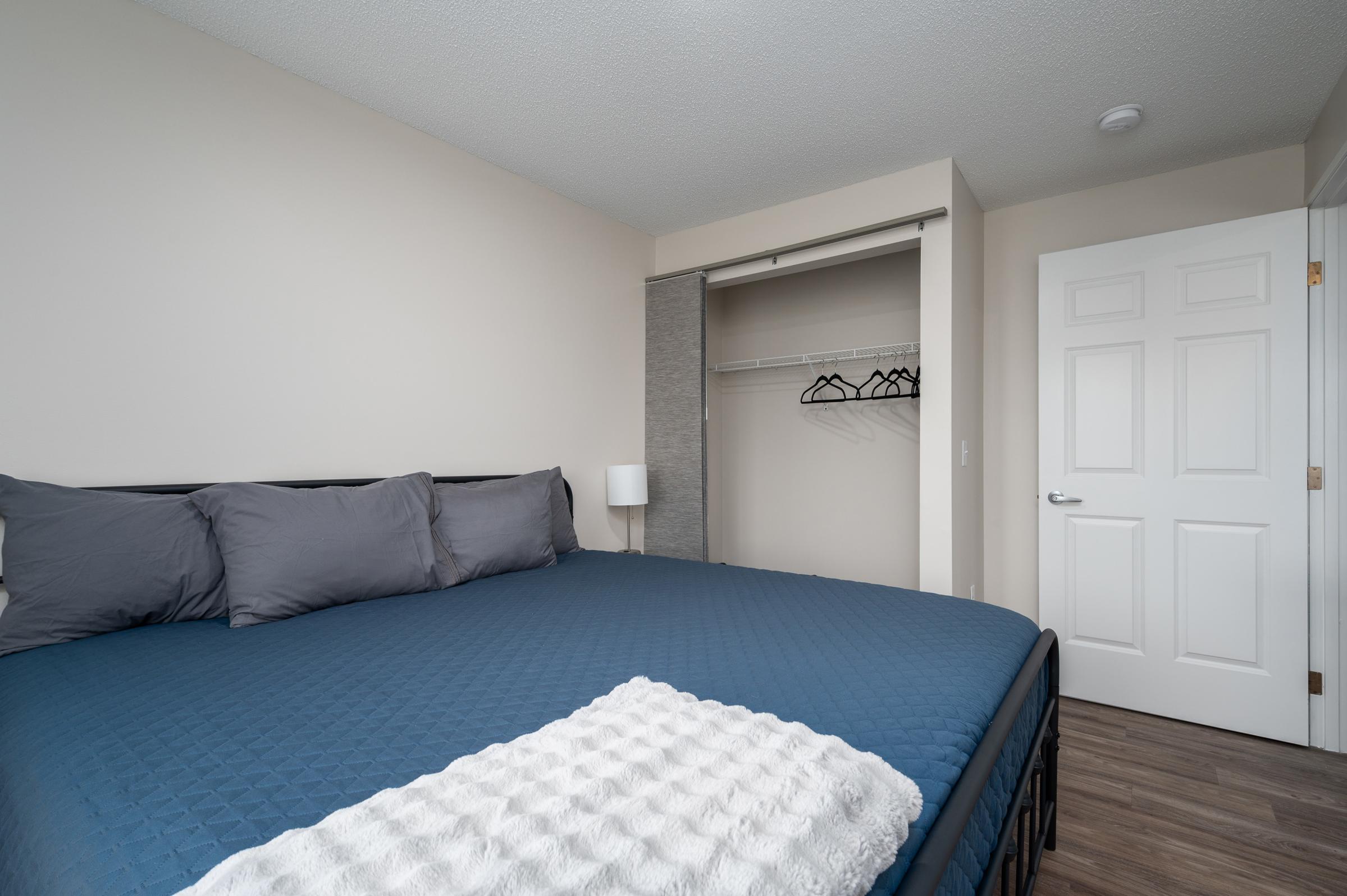 A cozy bedroom featuring a large bed with blue bedding and gray pillows. There is a white throw blanket on the bed. A closet with hangers is visible, and a white lamp stands next to the bed. The walls are painted in a light color, creating a warm and inviting atmosphere. A closed door is seen in the background.