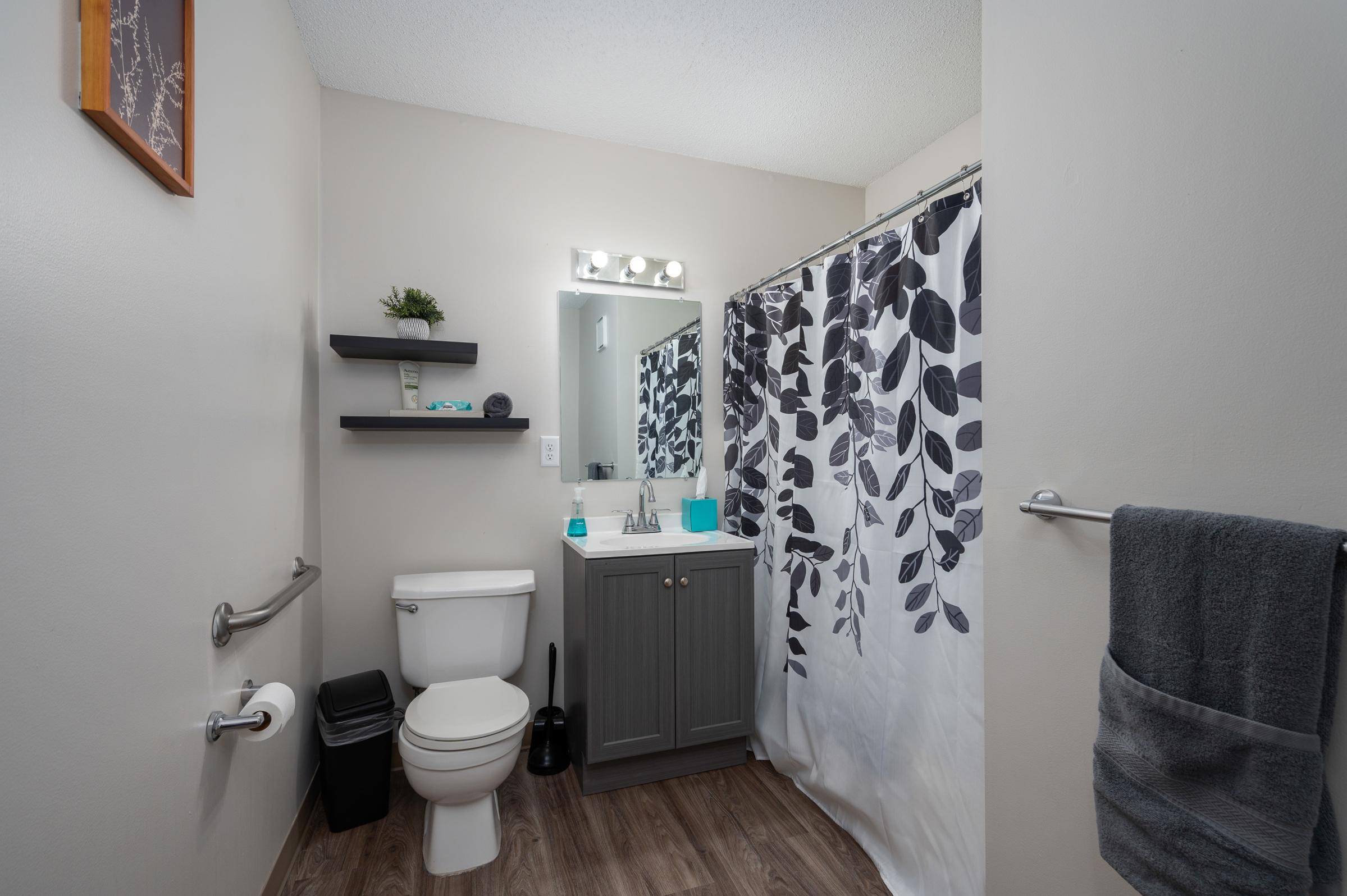 A modern bathroom featuring a white toilet, a gray vanity with a sink, a wall mirror, and light fixtures. The space is decorated with a black and white leaf-patterned shower curtain, a shelf with greenery, and a towel rack with a gray towel. The floor is wood-textured, and a black wastebasket is visible.