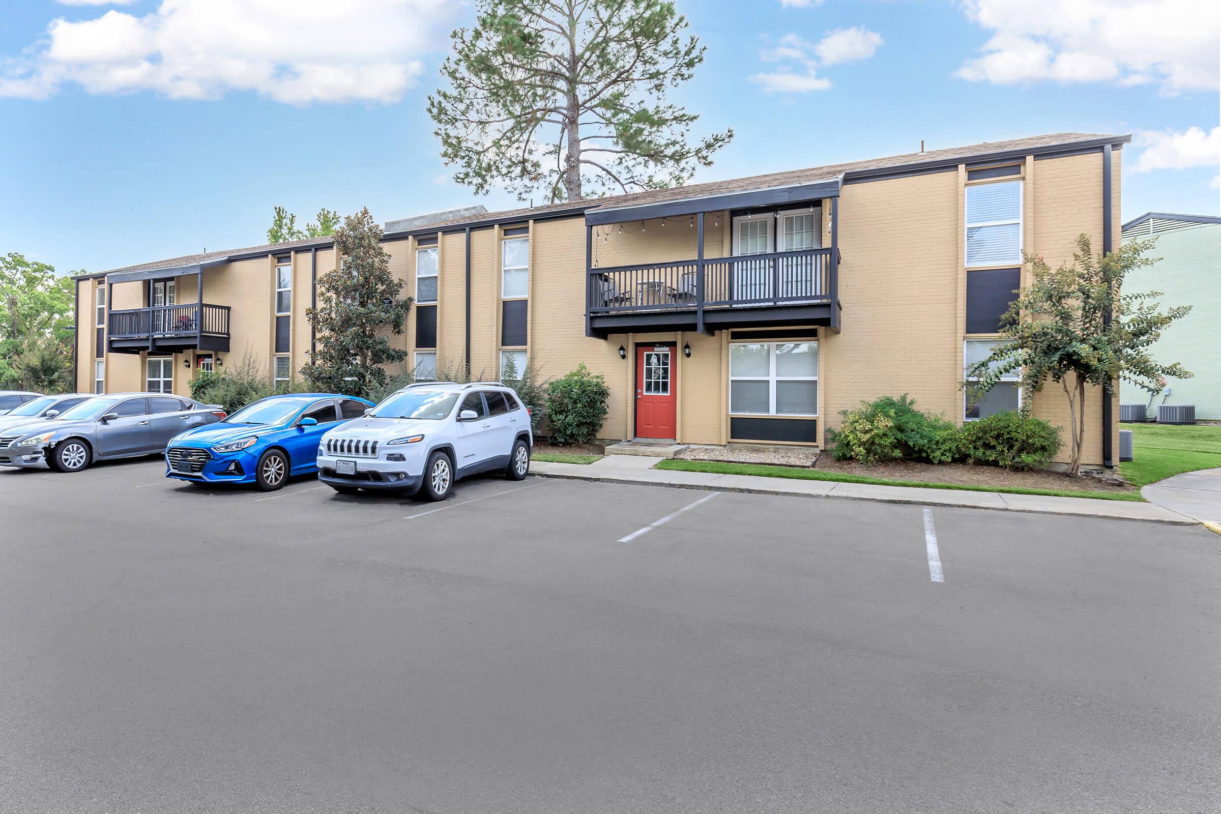 Two-story apartment building with a light brown exterior and balconies on the upper floor. There are several parked cars in front, including a silver SUV and a blue sedan. The lawn is green with small trees and shrubs, and the sky above is partly cloudy.