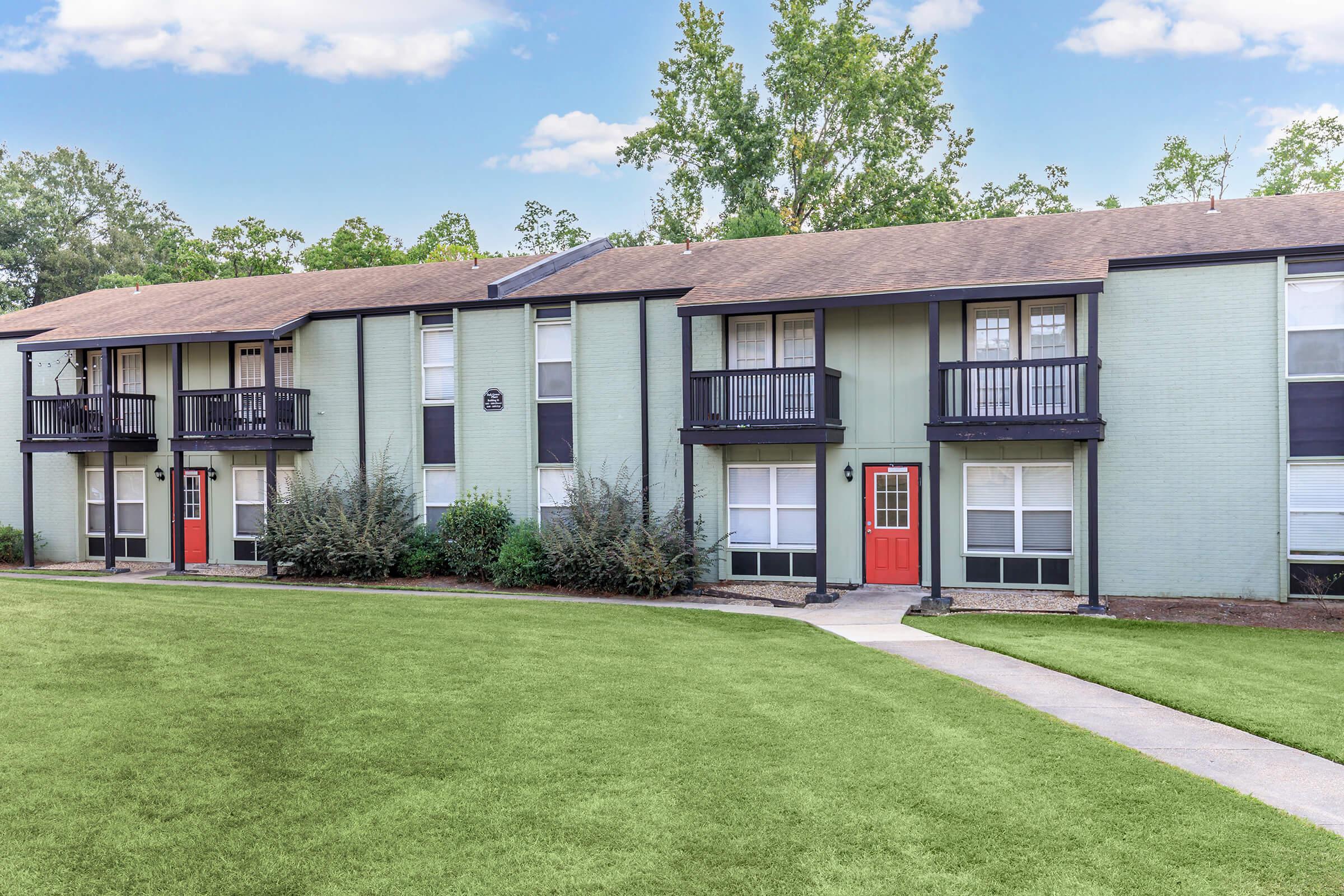Two-story apartment building with green siding, each unit featuring a small balcony and varying door colors (orange and red). The expansive lawn in front has manicured grass, and the background includes trees and a blue sky with scattered clouds. Pathway leads up to the entrance.