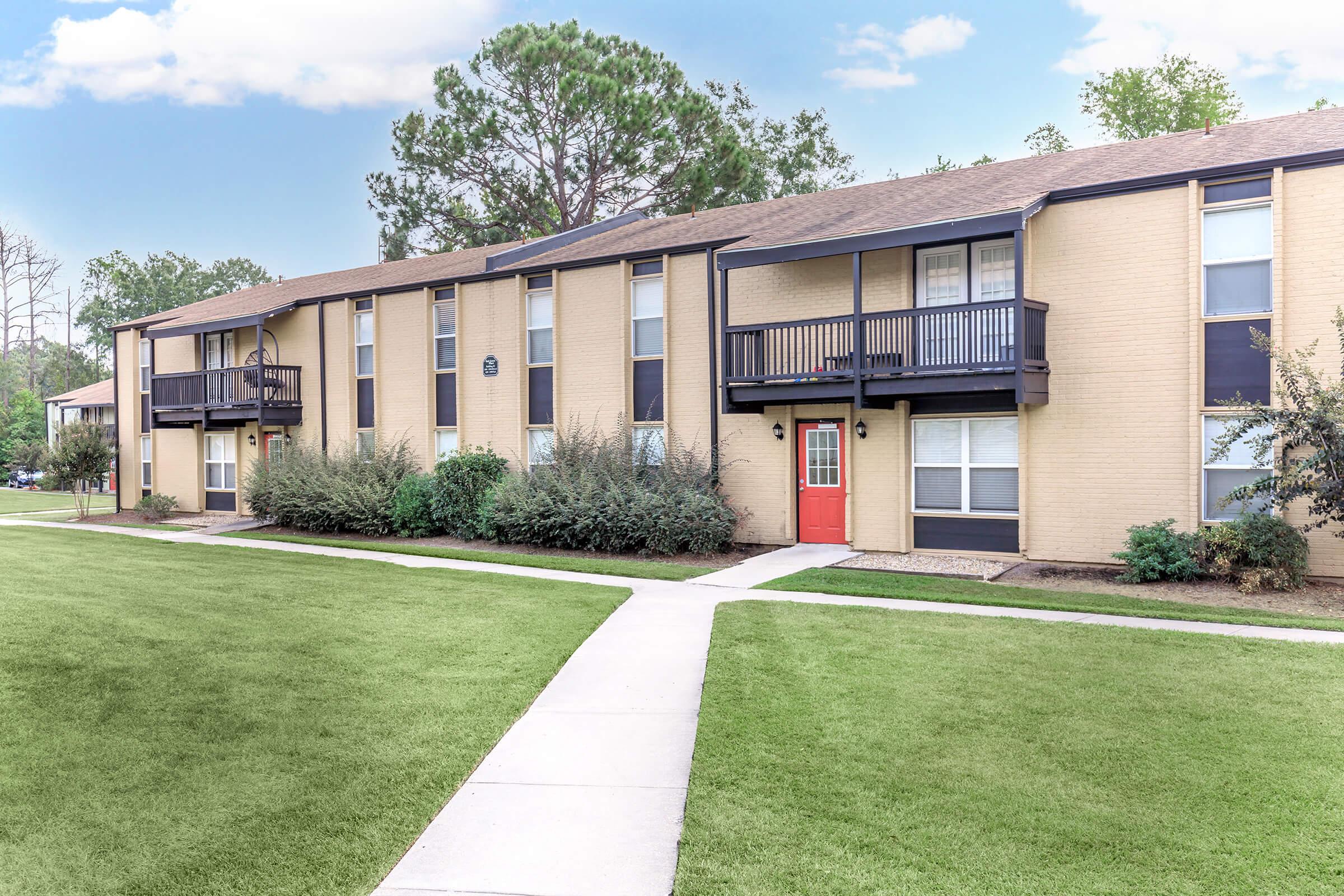 A two-story apartment building with a brick facade, featuring multiple balconies. The central entrance has a bright red door, surrounded by landscaped grass and shrubs. Sidewalks lead up to the building, and trees are visible in the background under a partly cloudy sky.