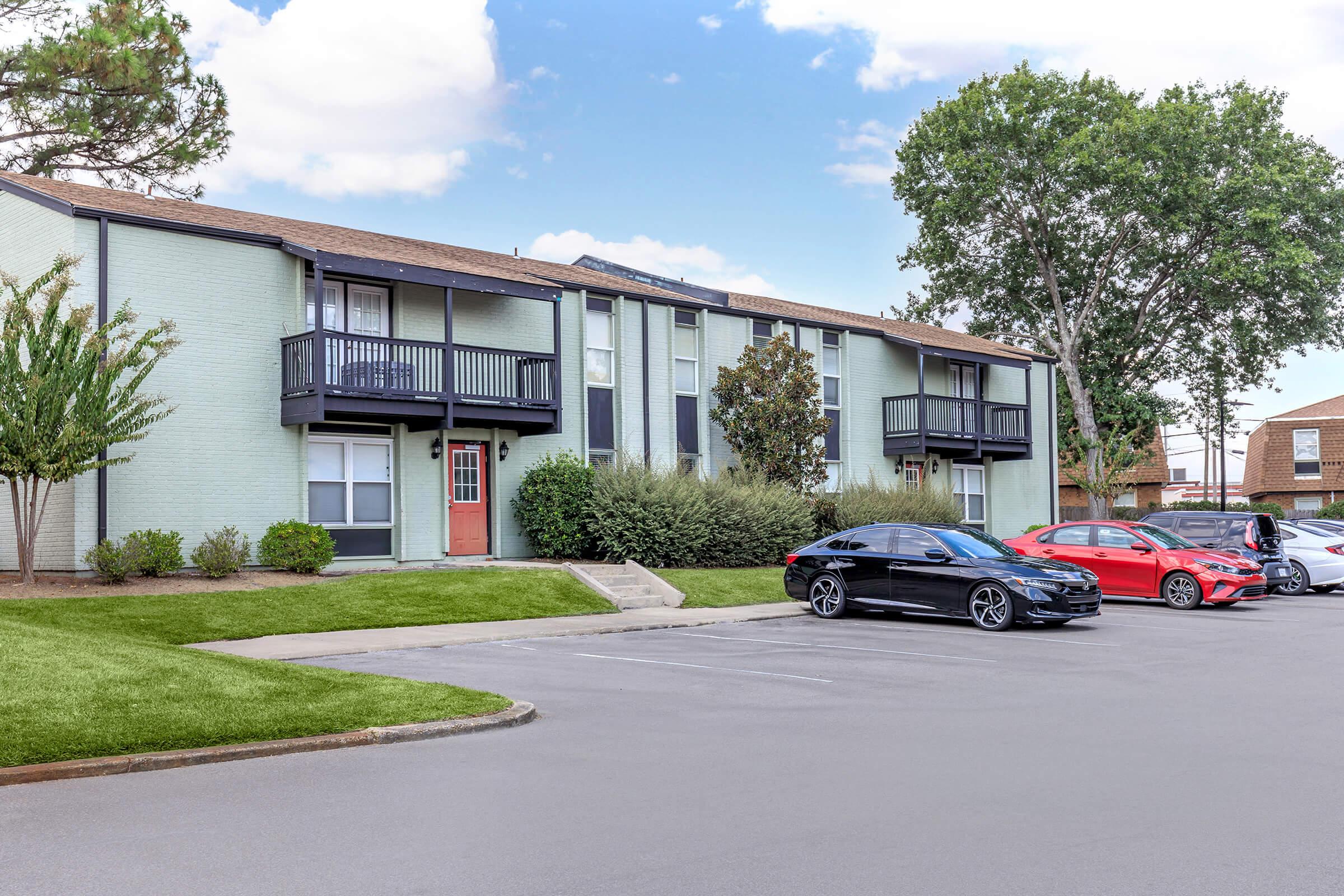 A row of multi-unit apartment buildings with green exteriors. The buildings have balconies and a central staircase. In front, there are two parked cars: a black sedan and a red sports car, on a paved area surrounded by grass and trees. The sky is partly cloudy.