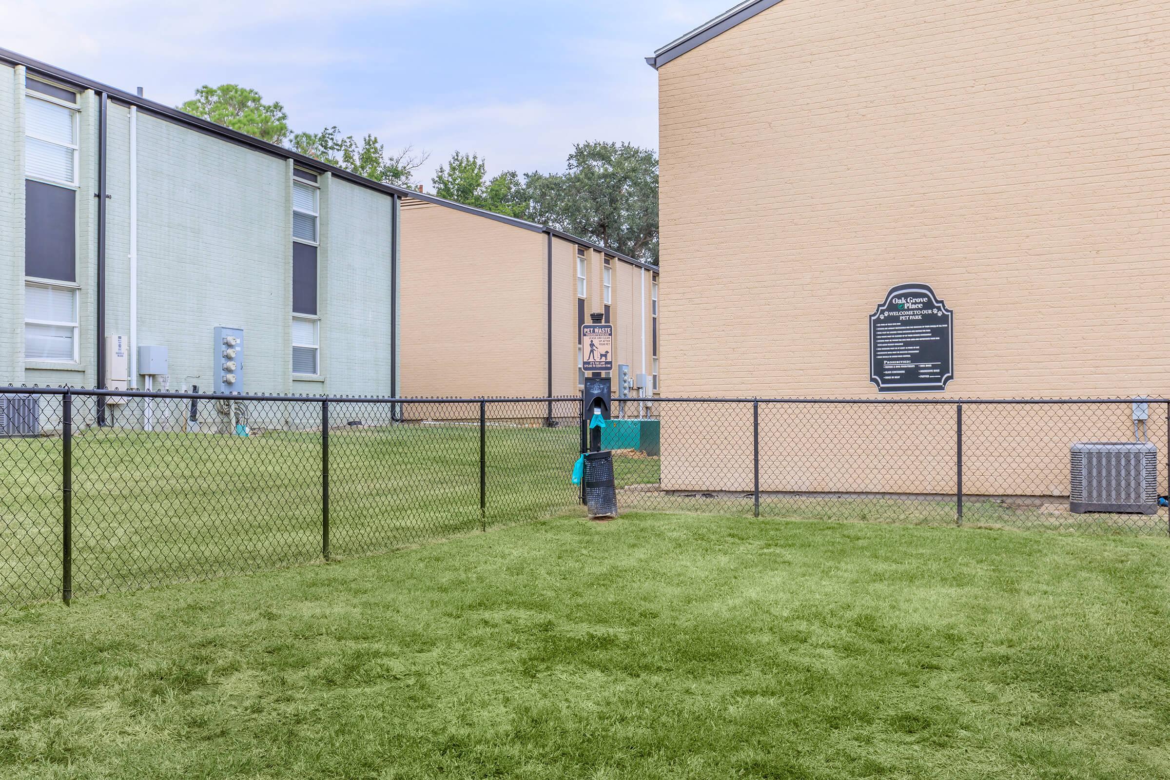 A fenced grassy area between two apartment buildings, with a sign attached to one building. A person dressed in blue is seen in the background. The setting appears to be residential, featuring well-maintained grass and utility structures.