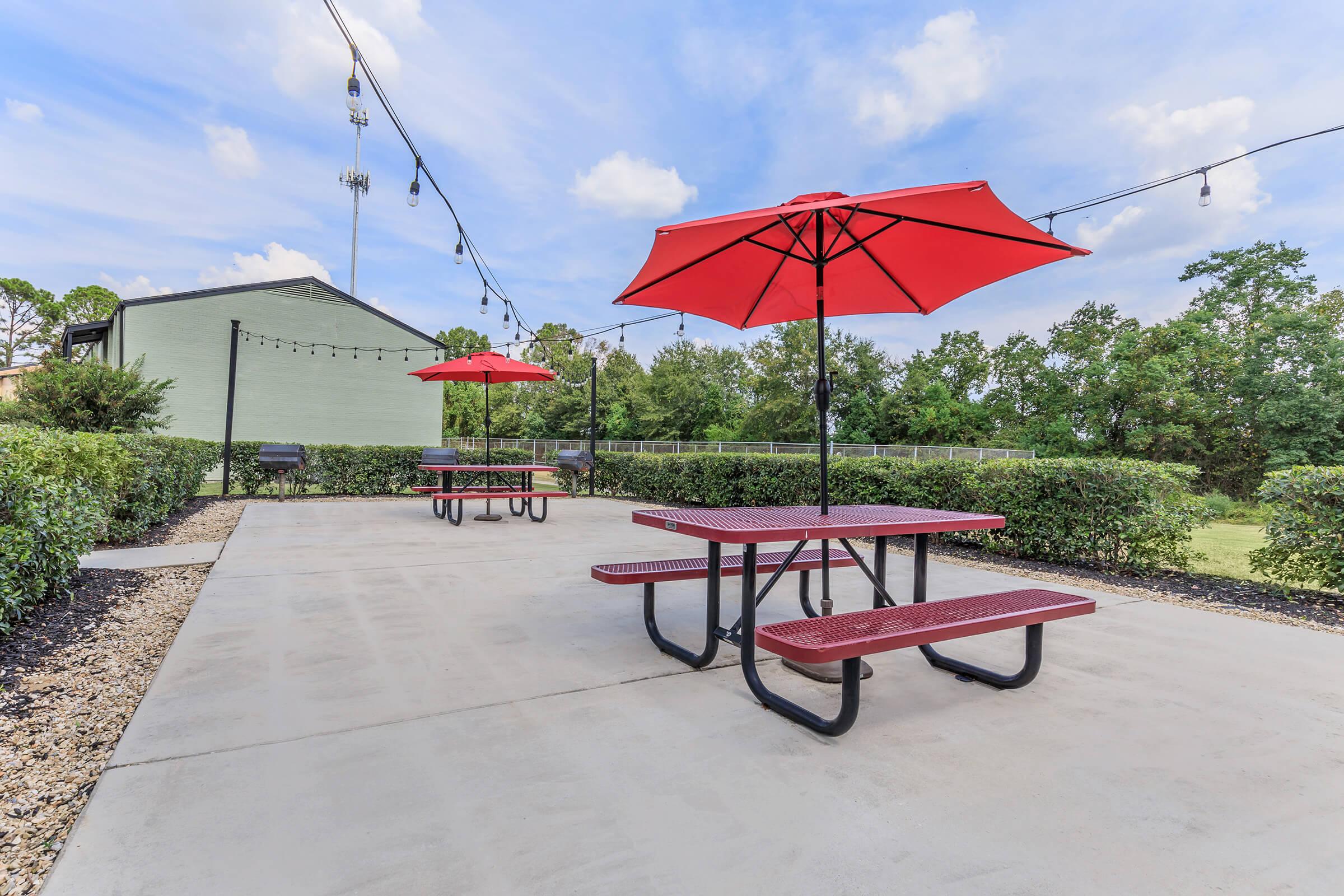 Outdoor patio area featuring two red picnic tables with umbrellas. The concrete surface is surrounded by green shrubbery, and there are string lights overhead. In the background, there's a light green building and trees under a partly cloudy sky.