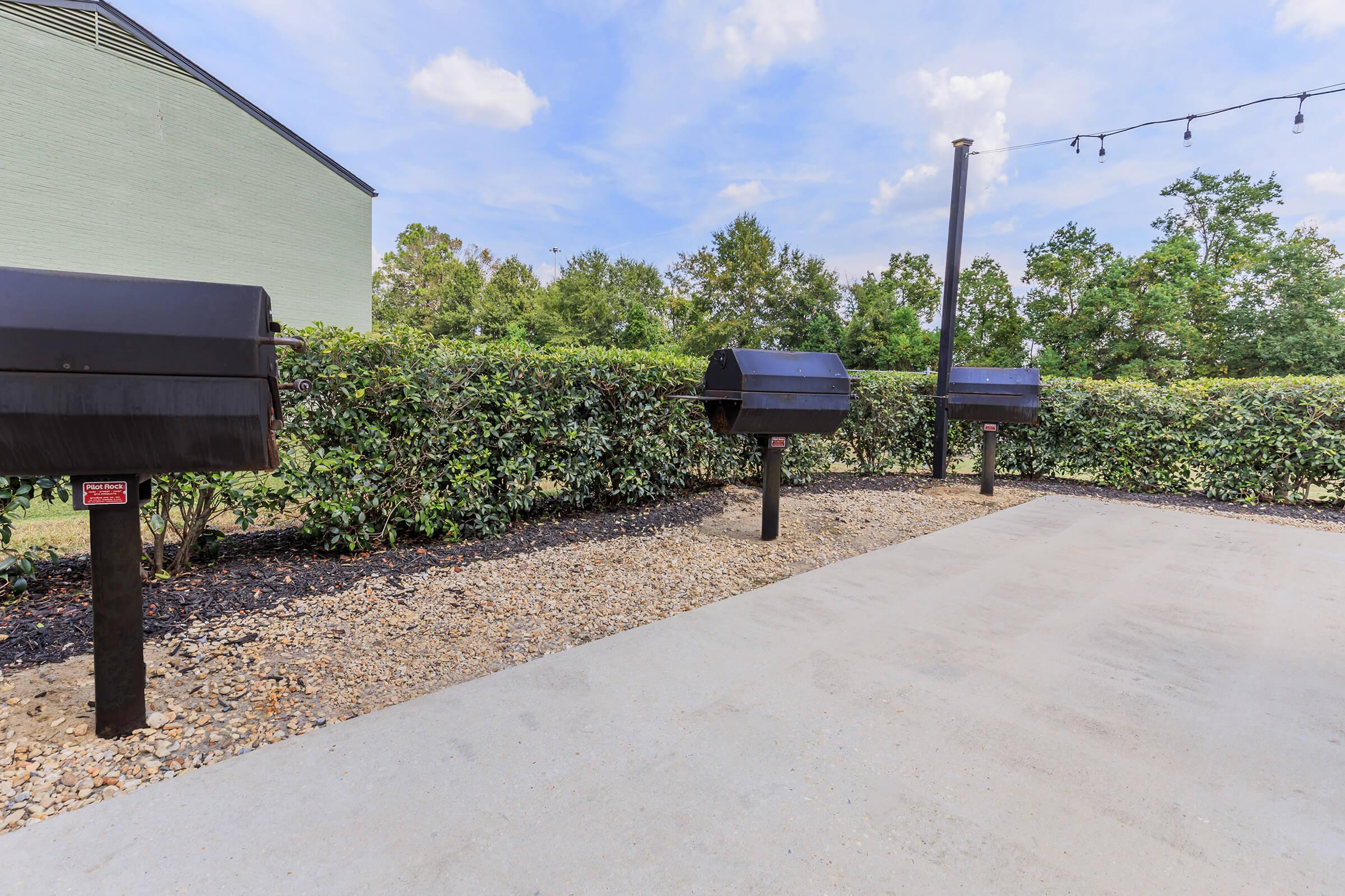 A landscaped outdoor area featuring three black charcoal grills positioned on pedestals, surrounded by neatly trimmed hedges and a gravel pathway. The background includes green trees and a blue sky with a few clouds, creating a pleasant atmosphere for outdoor cooking and gatherings.