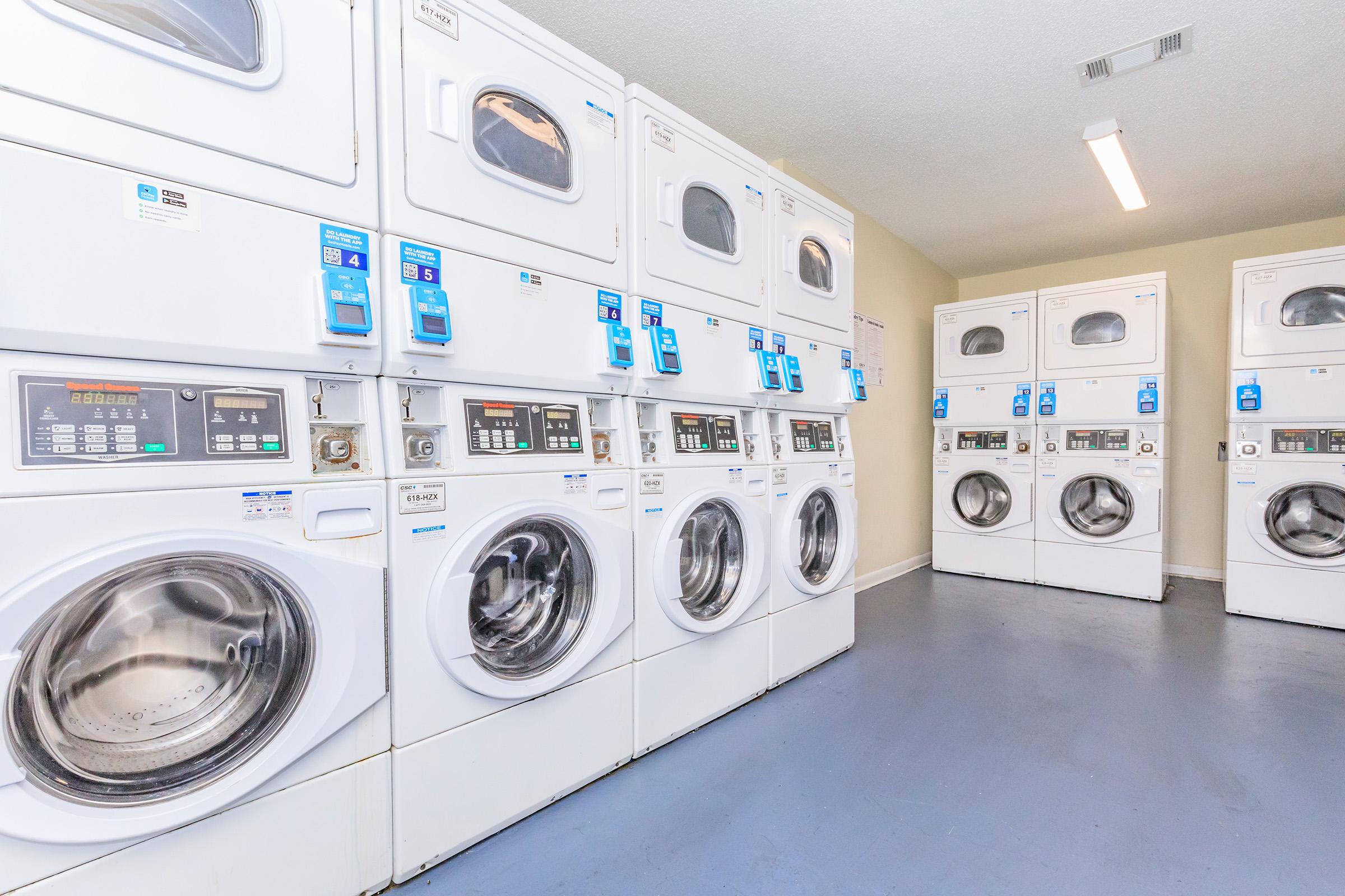 A clean laundry room with several stacked washers and dryers. The machines are white, with control panels featuring digital displays and coin slots. The floor is gray, and the walls are light-colored, creating a bright and organized atmosphere for laundry tasks.