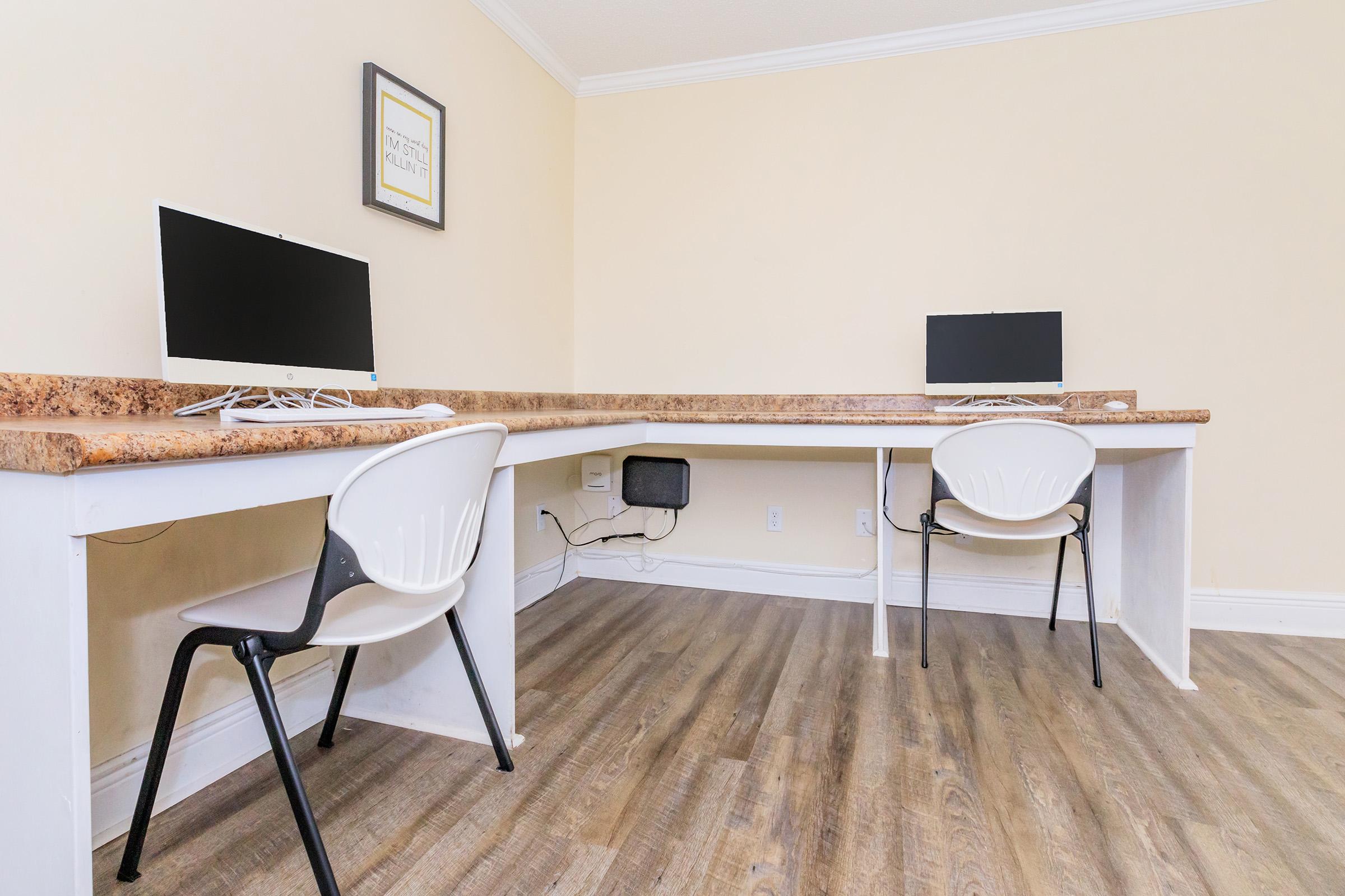 A minimalist office space with two white chairs positioned at a wooden desk. Each chair faces a desktop computer with black screens. The walls are painted a light color, and there is a framed picture on the wall. The flooring appears to be a wood-like laminate.