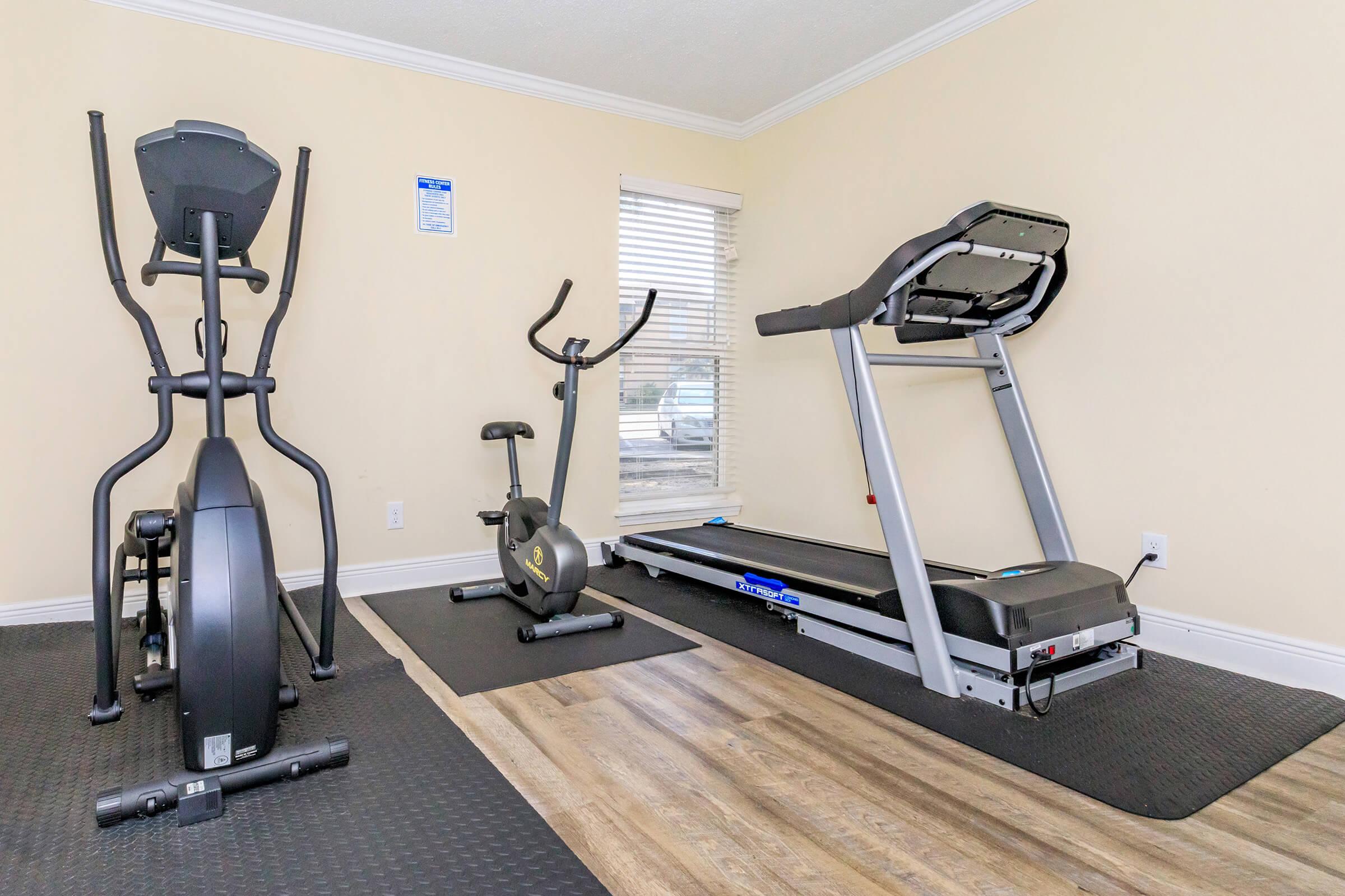 A fitness room featuring an elliptical machine, a stationary bike, and a treadmill, all positioned on black mats. The walls are painted a light color, and there is a window with blinds allowing natural light to enter. The flooring has a wood-like appearance.