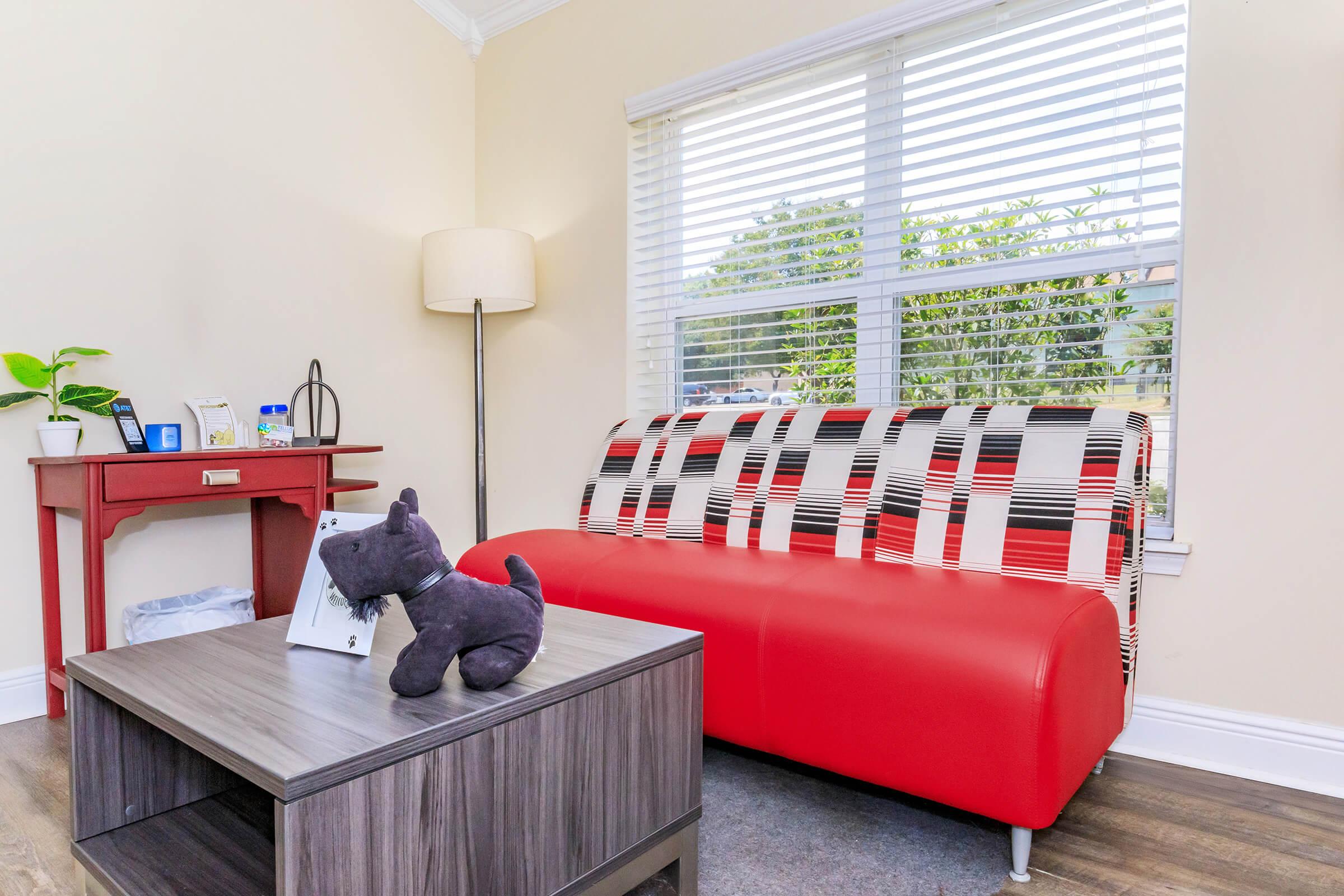 A modern living space featuring a red and black patterned couch, a wooden coffee table with a black cat figurine on top, and a small side table with decorative items. Natural light streams in through a window adorned with blinds, revealing greenery outside. The walls are painted a warm beige color.