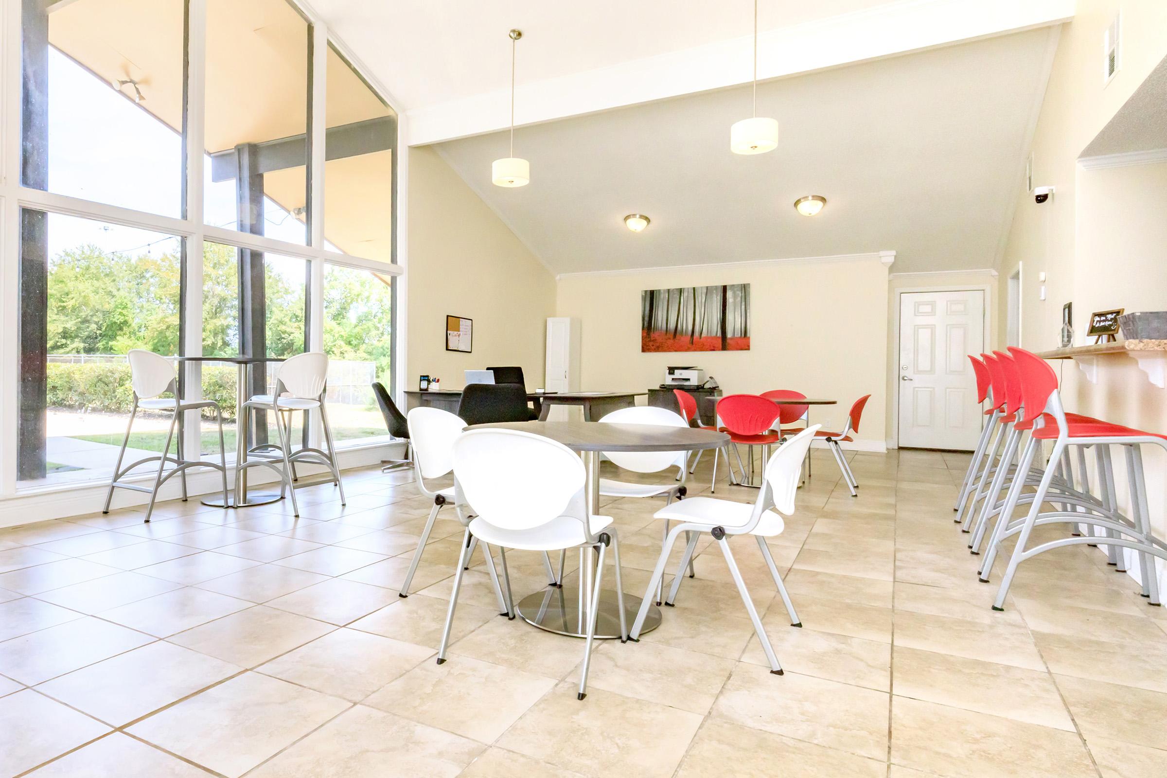 A spacious and modern communal space featuring tables and chairs with a mix of red and white seating. Large windows allow natural light to fill the room, providing a view of greenery outside. There is a printer and a bulletin board on the wall, adding to the functional atmosphere.