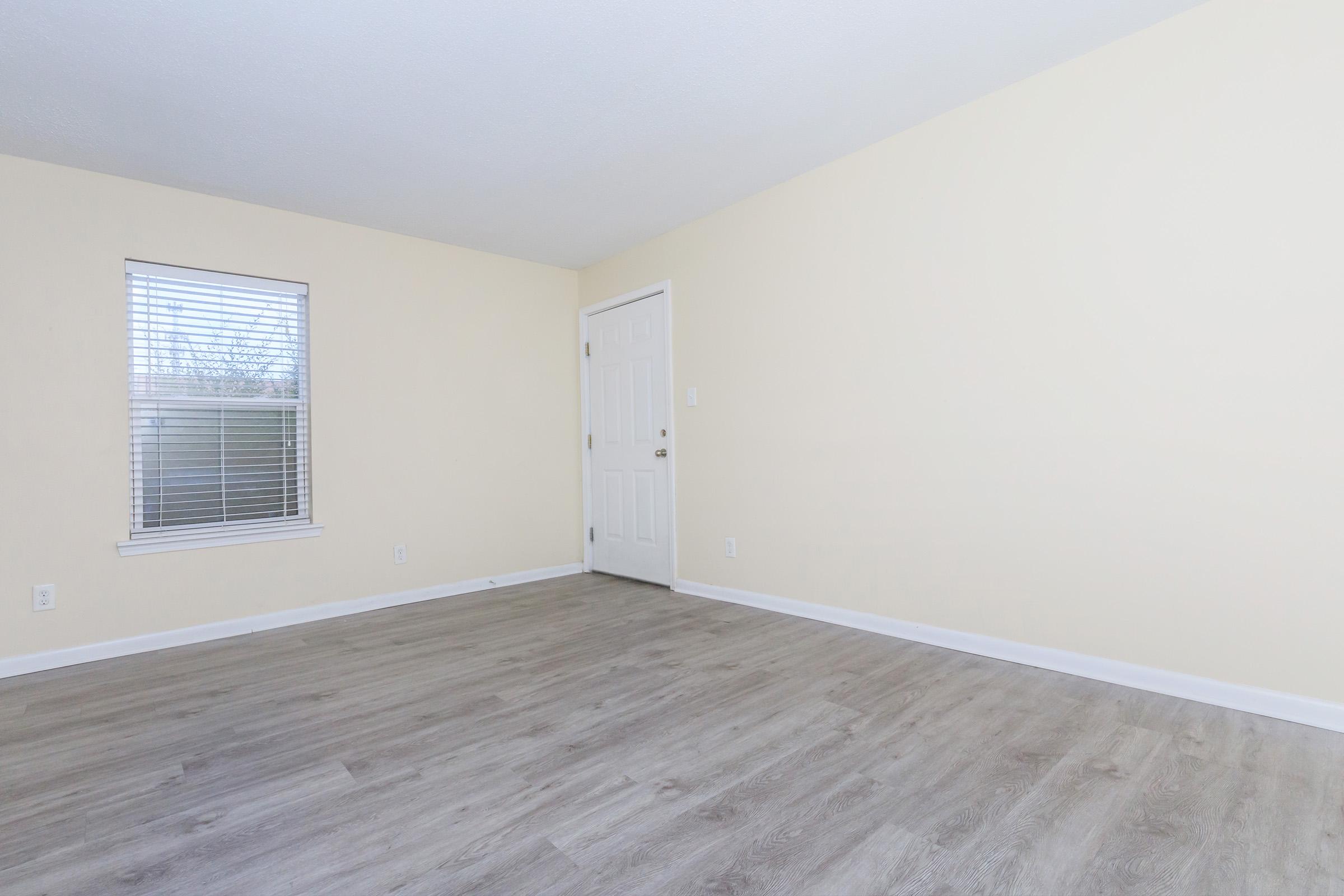 A spacious room with light yellow walls, a window with white blinds, and a door. The floor is made of light-colored wood laminate. The room is empty and well-lit, creating a bright and airy atmosphere.